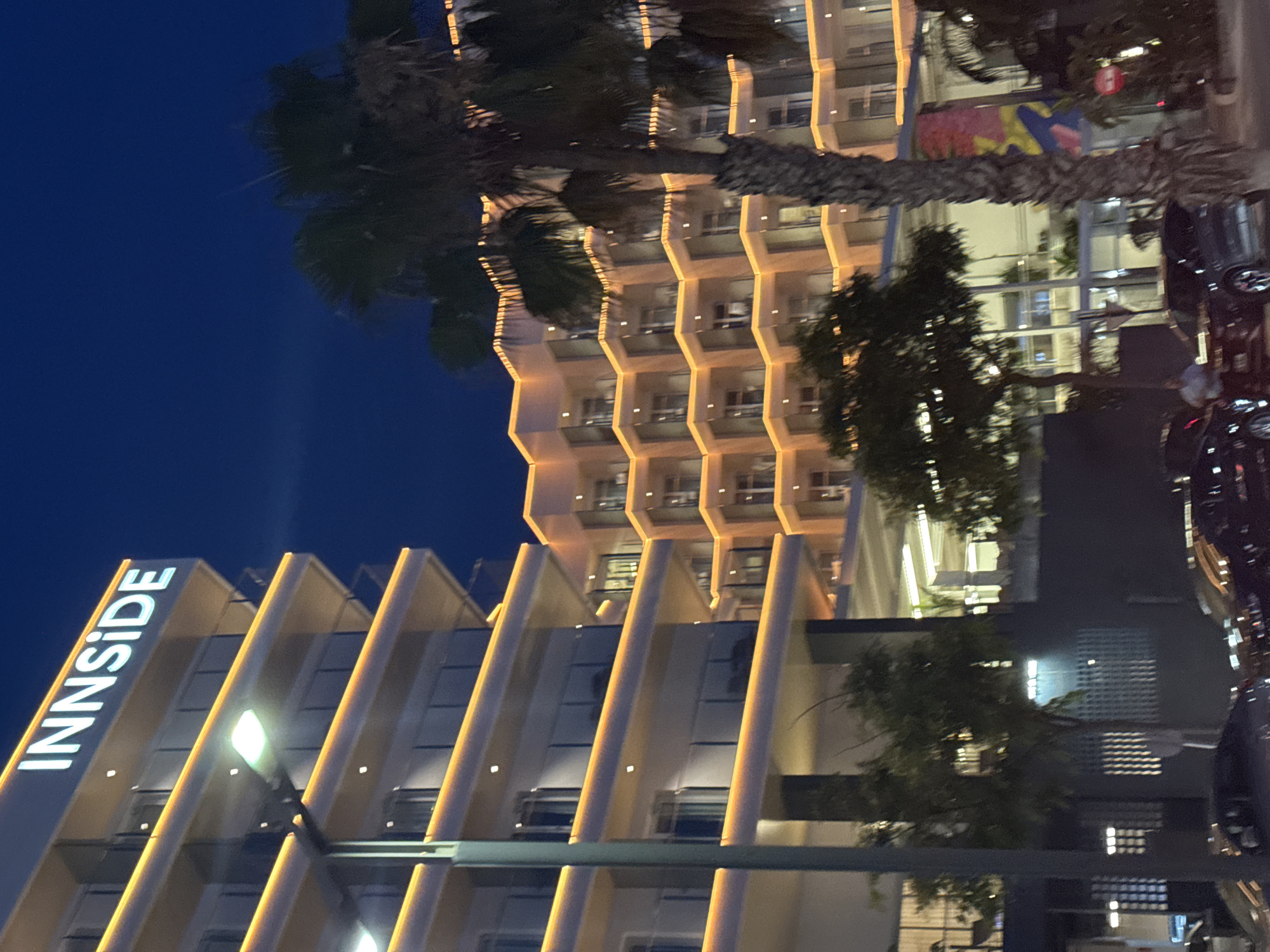 The image shows a nighttime view of the INNSIDE Alicante hotel, located in Alicante, Spain. The hotel is a multi-story building with illuminated balconies. In the foreground, several cars are parked. A palm tree stands to the right of the hotel, partially obscuring the view of a second, similar hotel building. The sky is dark blue, indicating it is nighttime.