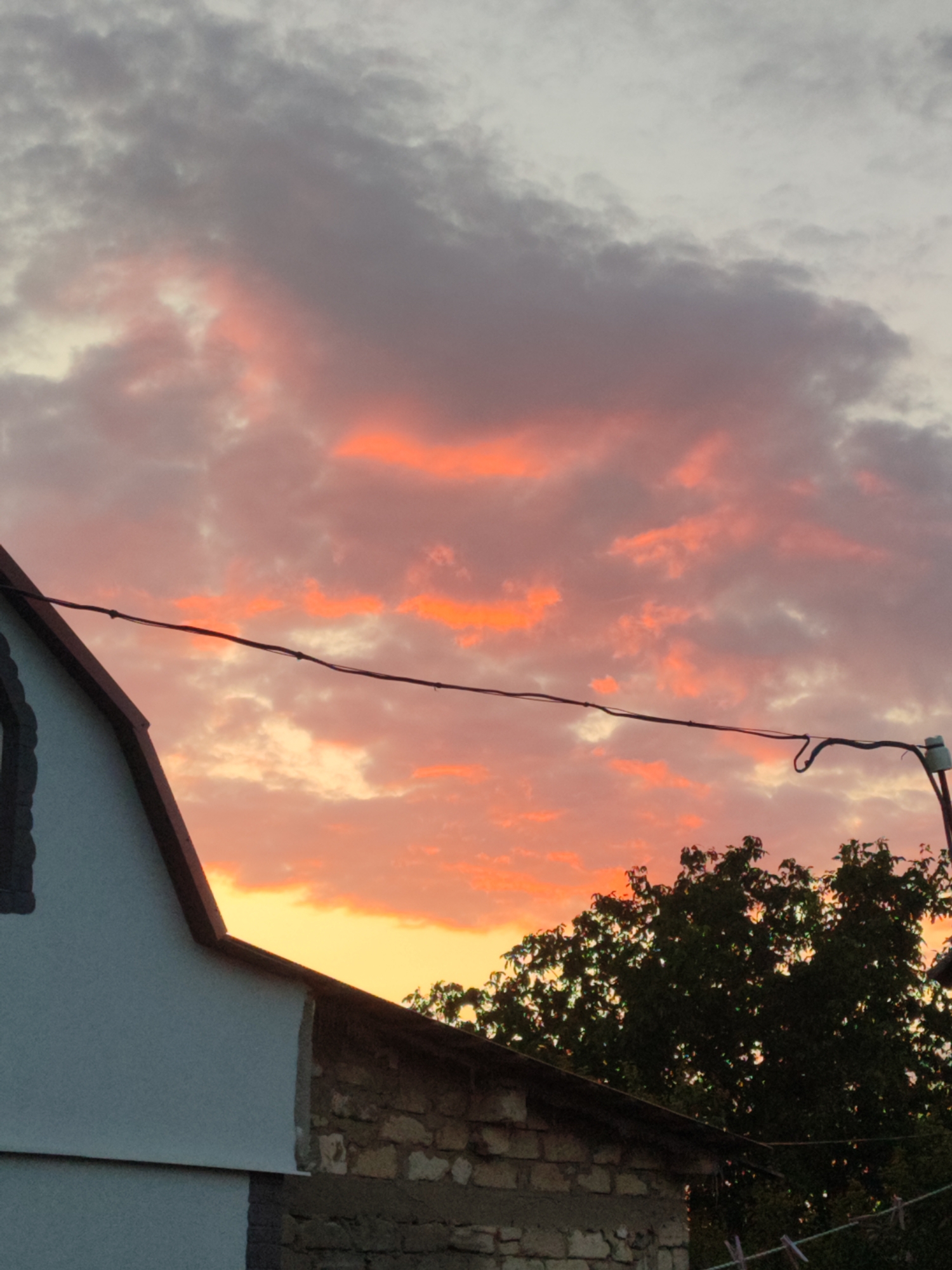 The image captures a sunset over Novosamarka, Ukraine, with vibrant hues of orange and pink illuminating the clouds. In the foreground, the roofline of a white-plastered building is visible, transitioning to exposed stone construction further along. A dark tree with dense foliage occupies the right side of the frame, partially obscuring the sky. Overhead, a thin black wire stretches across the scene, connected to a pole with an electrical insulator. The overall atmosphere is serene, with the dramatic sky being the focal point.