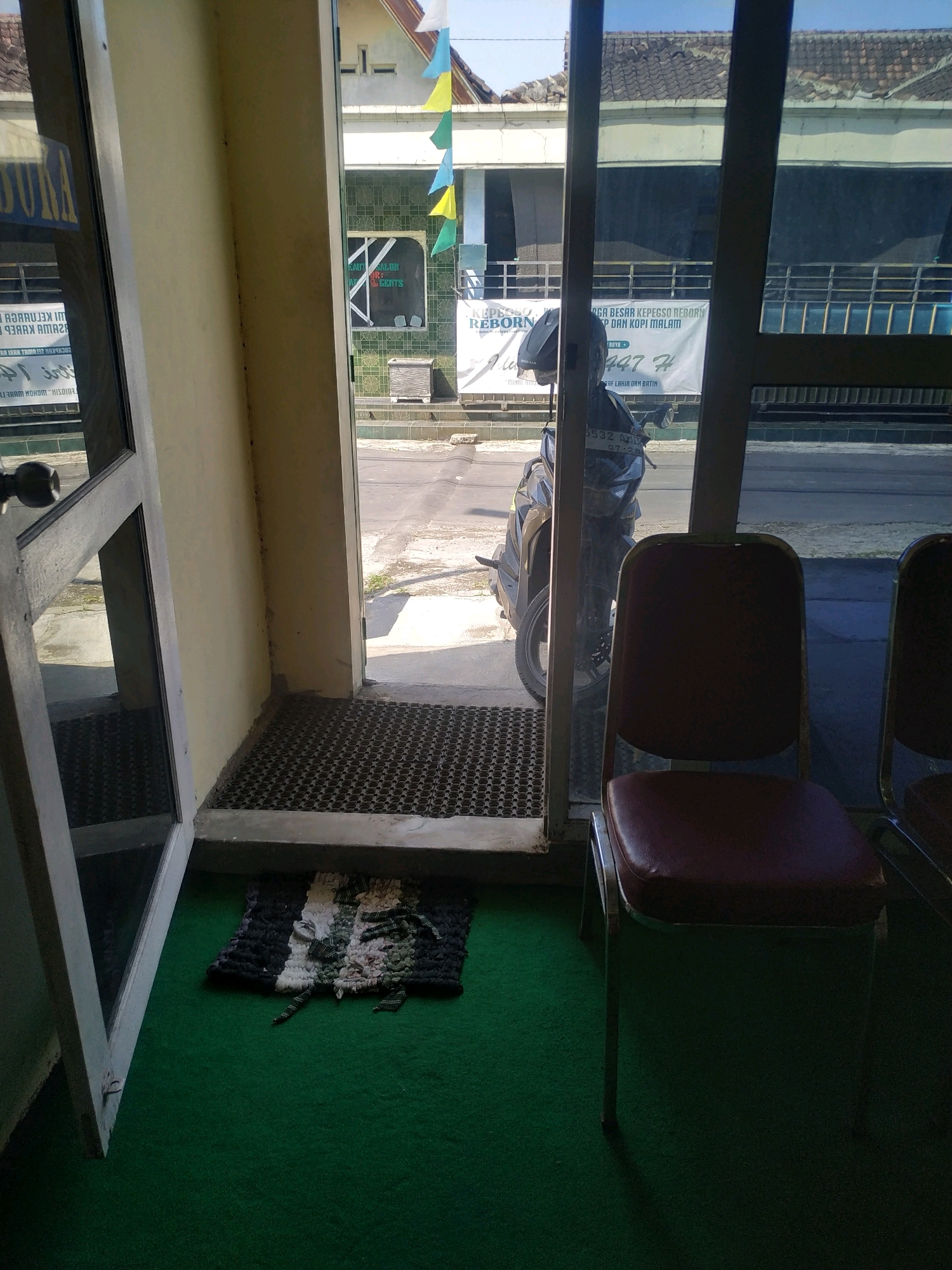 The image shows an interior view of a building with a doorway leading to an outdoor area. The doorway is to the left, with an open door revealing a mat and green carpeting inside. Through the doorway, a street is visible with a motorcycle parked on the side and a banner in the background. There are two chairs with dark red upholstery and chrome legs positioned inside the building, facing towards the street. One chair is fully visible, while the other is partially obscured. The exterior walls are light yellow, and the floor inside is covered in green carpet. The scene appears to be in Batan, Indonesia, as indicated by the presence of a motorcycle with Indonesian license plates. The lighting suggests it is daytime, with sunlight casting shadows.
