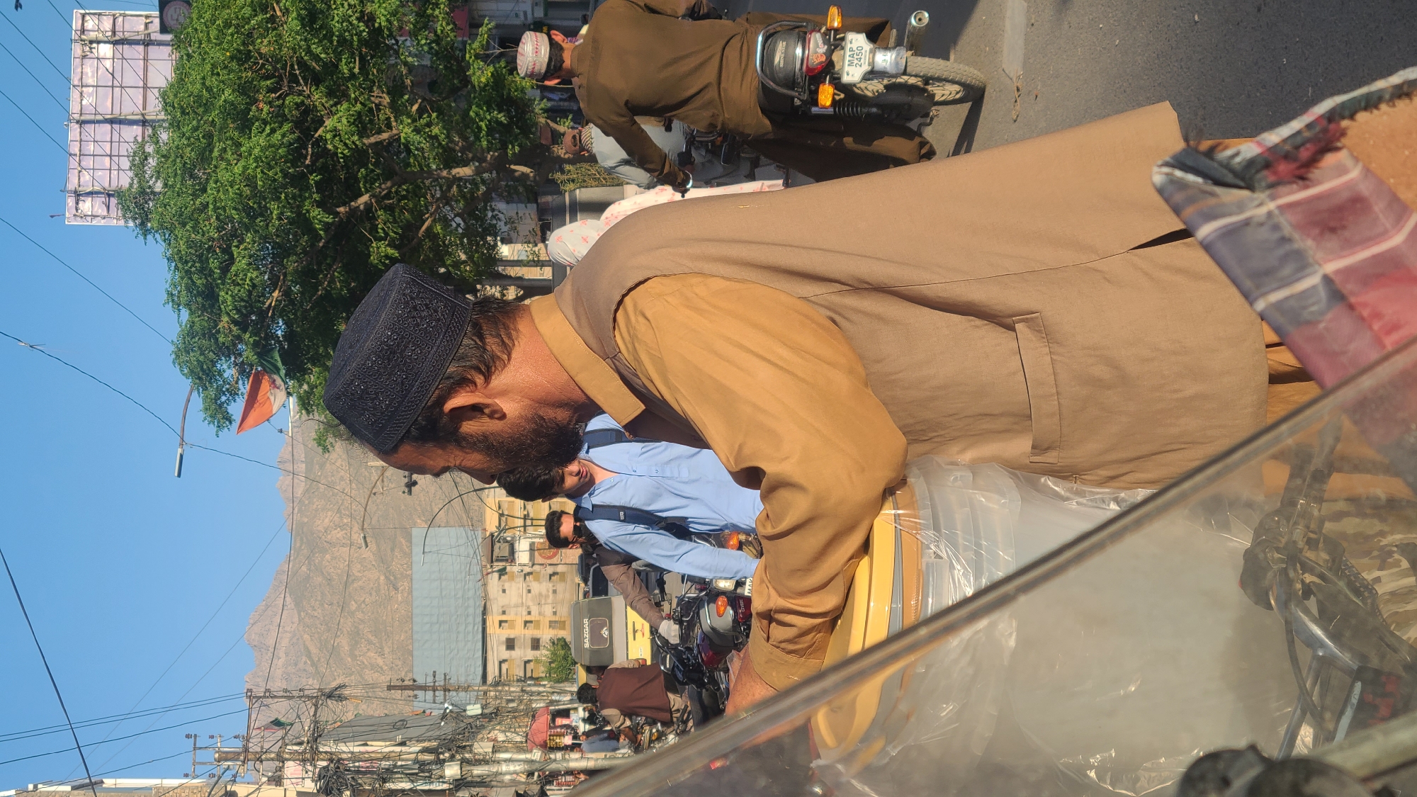 The image captures a vibrant street scene in Quetta City Tehsil, Pakistan, under a clear blue sky. In the foreground, a man wearing a light brown shalwar kameez and a dark, embroidered skullcap (kufi) is seen from the side. He has a beard and is leaning forward, seemingly attending to something within a transparent container or display, possibly from behind a vehicle's windshield, a part of which is visible in the lower right.
The midground features several other individuals engaged in daily activities. A man in a light blue shirt and a backpack is visible behind the foreground subject, and further back, another man is seated on a motorcycle. A motorcycle in the immediate background clearly displays a license plate with the text "MAP-2450". Most people are dressed in traditional South Asian attire.
The background reveals a bustling urban environment with buildings lining the street and a large, verdant tree prominent on the left. Numerous tangled power lines and utility cables crisscross the sky, a common feature in many urban areas of the region. Dominating the far background are majestic, rugged brown mountains, characteristic of the landscape surrounding Quetta. An orange and green flag is visible flying from a pole. The bright, clear daylight suggests the time is either morning or afternoon. The scene depicts a typical day in a busy Pakistani city, with a blend of human activity, urban infrastructure, and natural beauty.