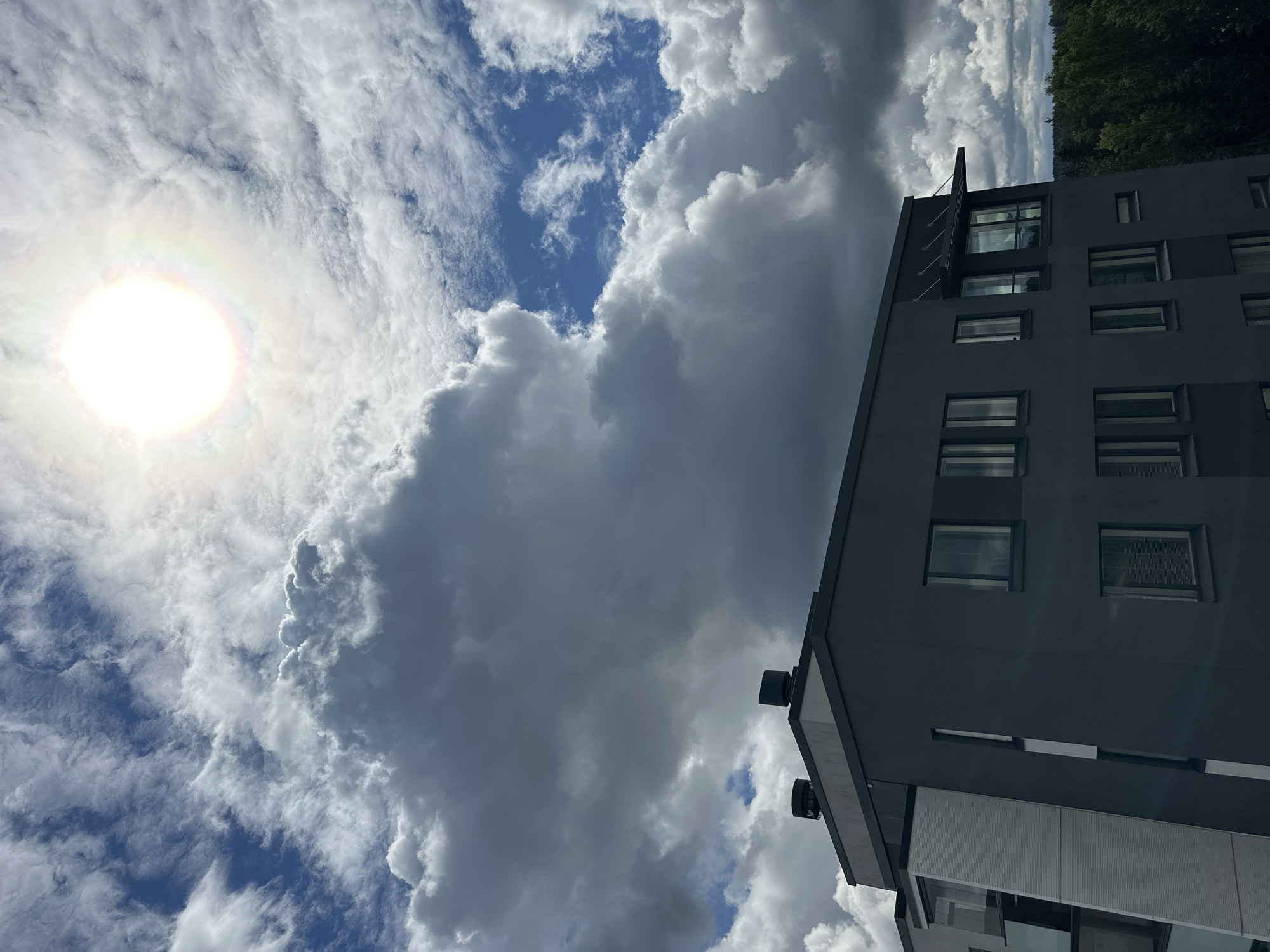 An exterior, eye-level, vertical, full shot captures a modern, dark gray apartment building against a dramatic sky filled with large, puffy, gray and white clouds and a bright sun with a halo effect. The building has a light gray facade on the left, with a sloped section featuring horizontal white siding. The main section of the building is dark gray with multiple windows, some reflecting the cloudy sky. Two dark cylindrical vents are visible on the roof. Beyond the building to the right, a dense, dark green forest stretches to the horizon under the cloudy sky. The sun is positioned in the upper right quadrant of the frame, partially obscured by clouds, casting a bright glow with visible prismatic colors. The lighting suggests it is daytime.