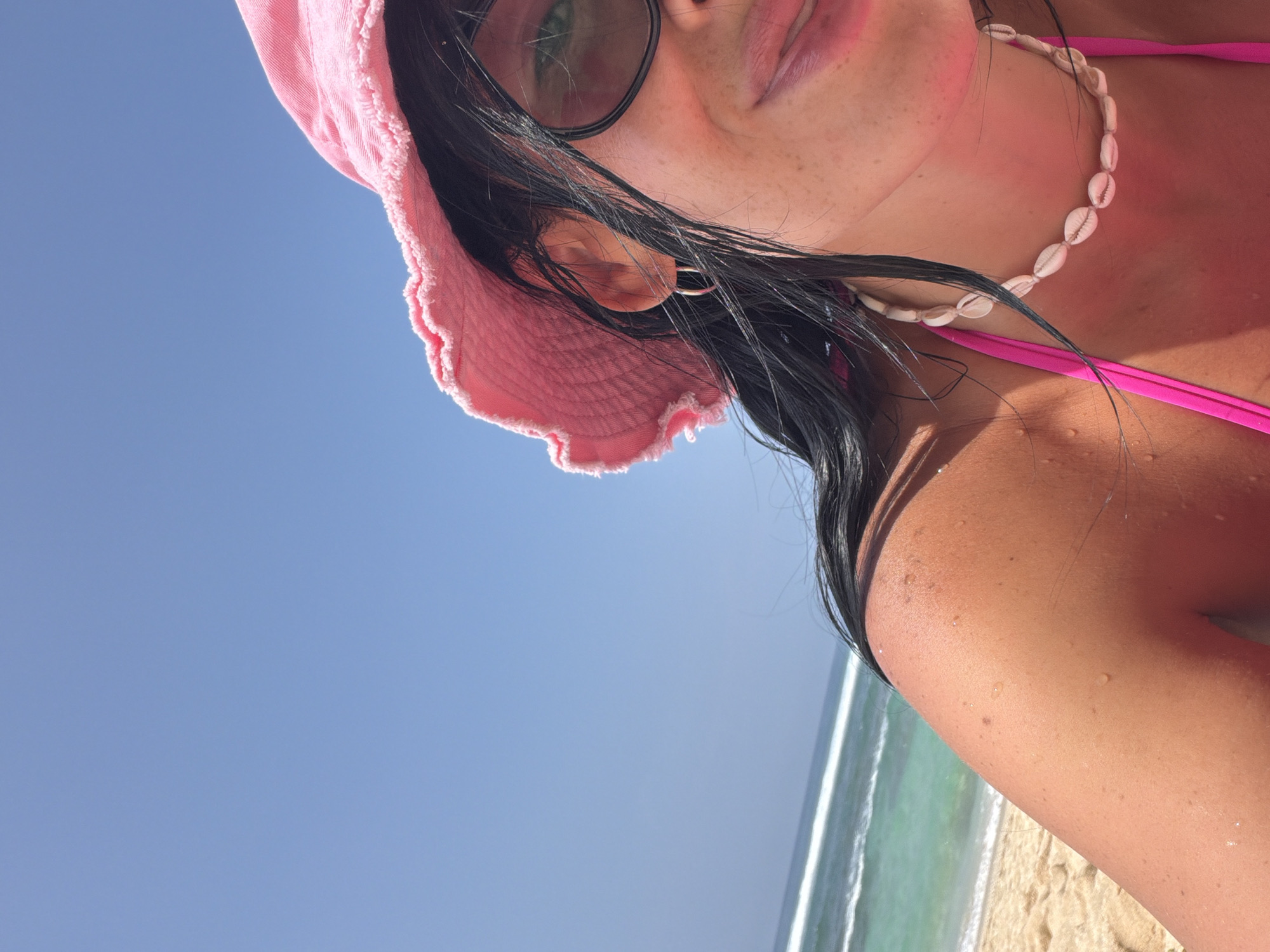 A close-up, upward-angled image captures a female individual on a beach in Ungasan, Indonesia. The subject is shown from the mid-chest upwards, with tanned skin and visible water droplets on her right shoulder. She wears a bright pink bucket hat with a frayed brim, dark sunglasses, a pink bikini top, and a necklace composed of numerous off-white cowrie shells. Her dark hair appears wet or windswept, and a small, silver-colored hoop earring is visible on her right ear. The lower right portion of her face is visible, showing a faint smile. The background features a clear blue sky in the upper left quadrant, transitioning to turquoise ocean water with white breaking waves in the mid-left, and tan sand along the lower left edge. The scene is brightly illuminated by natural daylight.