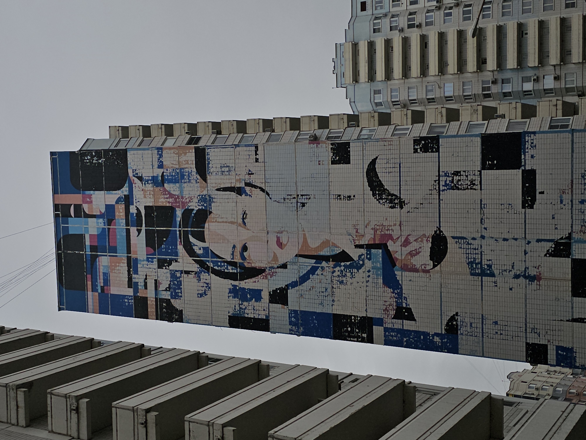 A large mural covers the side of a tall apartment building in Odintsovo, Russia. The mural depicts a stylized portrait of a woman's face, rendered in a pixelated style with geometric shapes and abstract elements. The woman has dark hair and is looking to her right. The mural is composed of various colors, including blue, pink, black, and white.
The apartment building itself is grey and features numerous balconies. To the left of the building with the mural, another apartment building with similar balconies is visible. To the right, another, shorter apartment building with a light blue and grey facade is partially visible. The sky above is overcast and grey. Power lines are visible against the sky. The image appears to be taken from a low angle, looking up at the buildings.