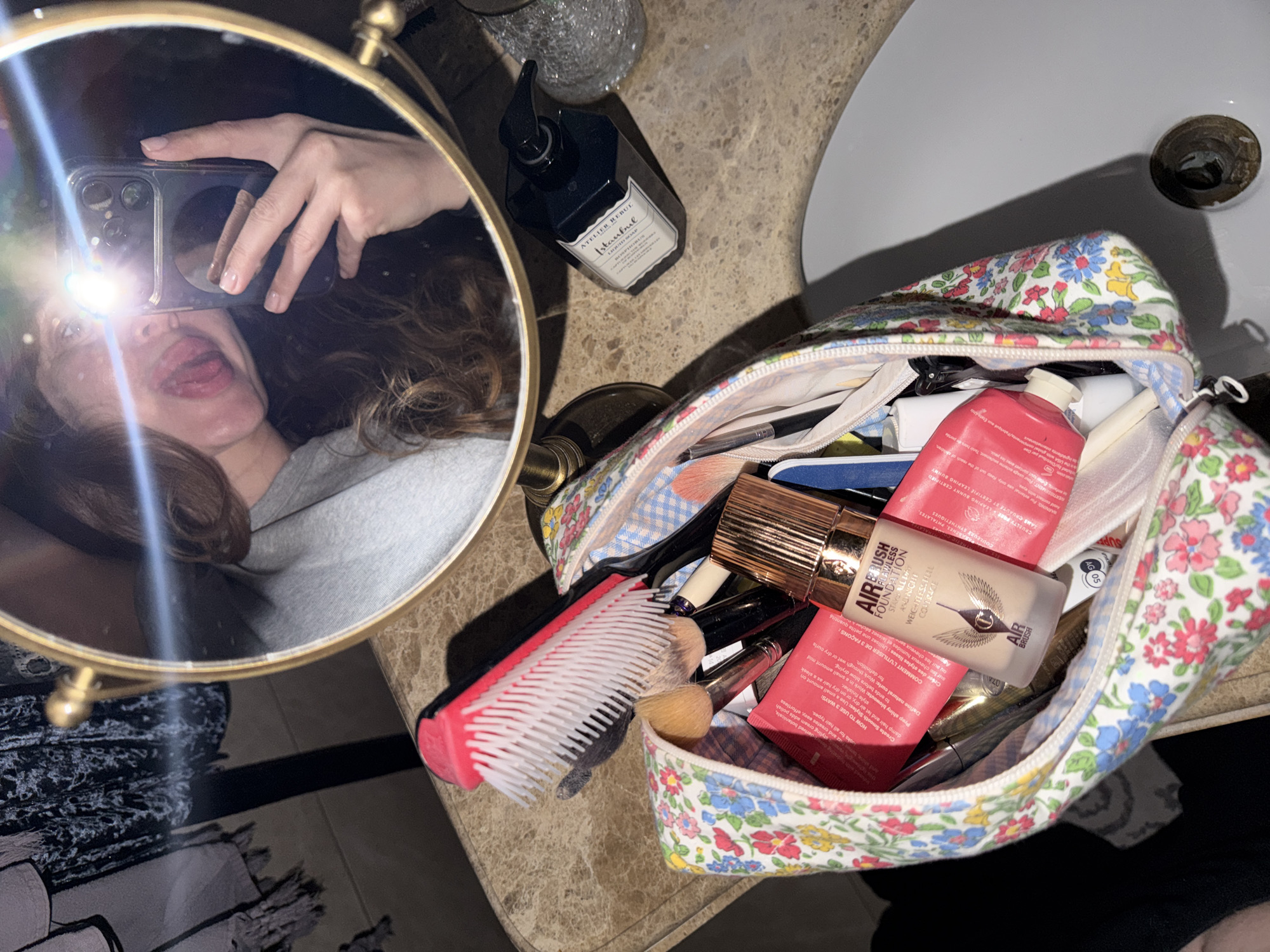 The image shows a bathroom scene featuring a woman taking a selfie in a round mirror. Her hair is curly, and she is sticking out her tongue. In the foreground, a makeup bag with a floral pattern is open, revealing various cosmetic items, including "Airbrush Flawless Foundation" and red tubes. A comb and makeup brushes are next to the bag. A soap dispenser is in the background, and the sink is visible on the right side of the frame. The location appears to be a bathroom with a marble countertop. There is no information indicating that this image was taken in the city of Mishutkino, Russia.