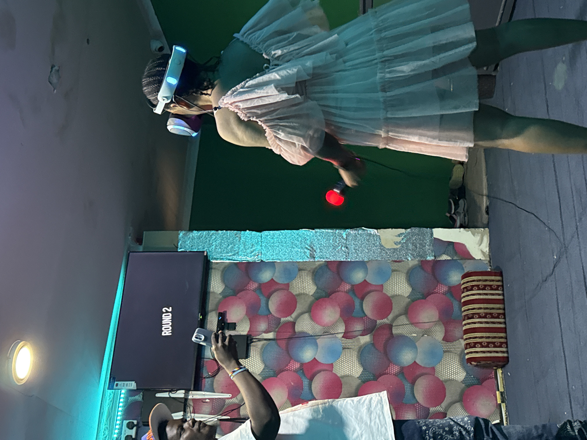 In Dar es Salaam, Tanzania, two individuals are engaged in a virtual reality experience in a dimly lit room. On the left, a person wearing a white t-shirt and a hat holds a smartphone. In the center, a large television screen displays "ROUND 2" in white text on a black background. To the right, a person wearing a pale pink off-the-shoulder dress and a VR headset is actively participating in the game, holding a red controller. The room features a wall with a circular patterned backdrop in shades of pink, blue, and white, and a long, striped cushion rests on the floor in front of it. The overall ambiance suggests a recreational setting for augmented or virtual reality gaming.