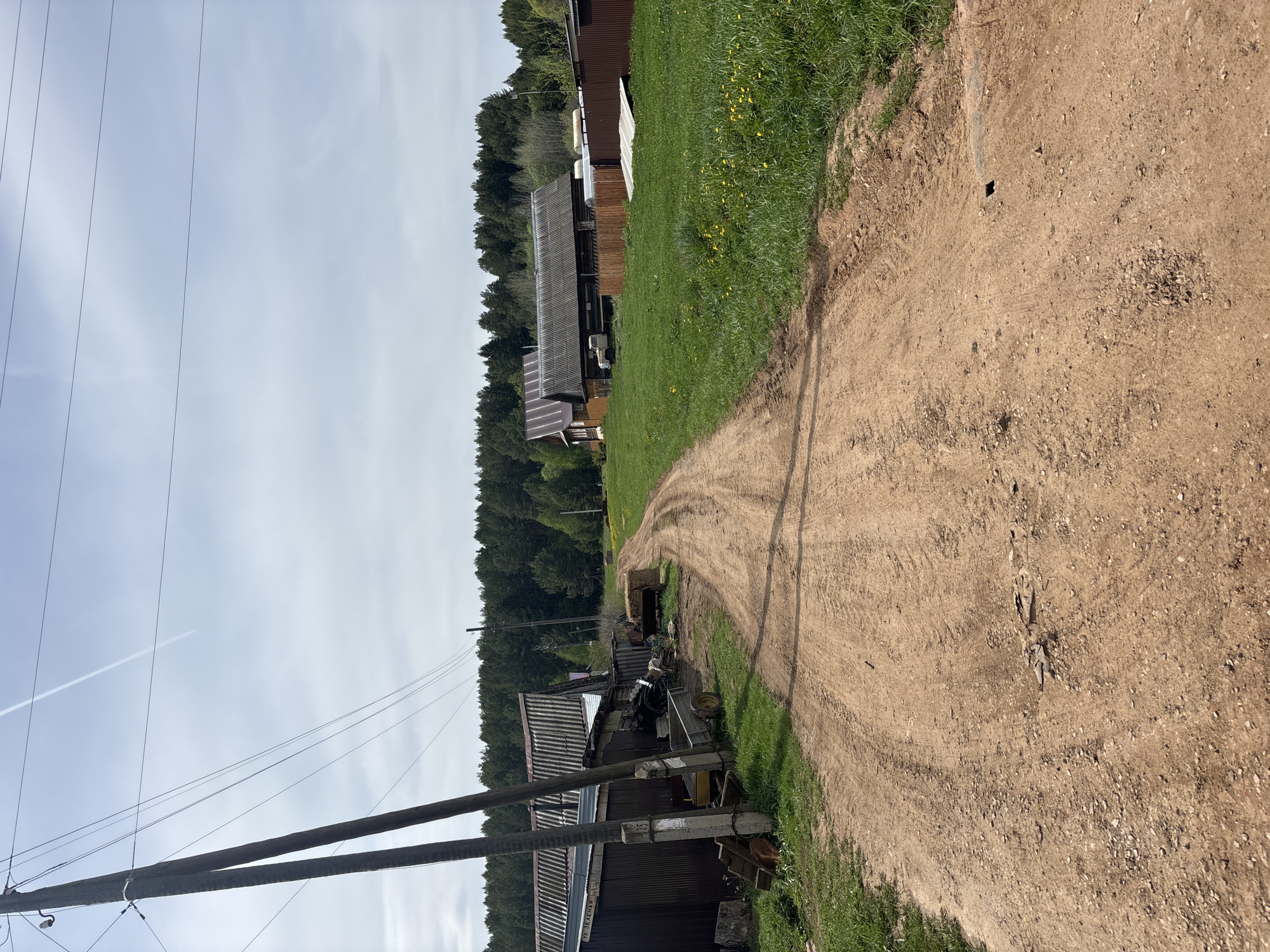 The image depicts a dirt road in Niva, Russia, flanked by grass and buildings. The road curves into the distance toward a forest. Power lines extend into the sky from a utility pole near a corrugated metal structure, cluttered with various objects. Aircraft contrails are visible in the sky.