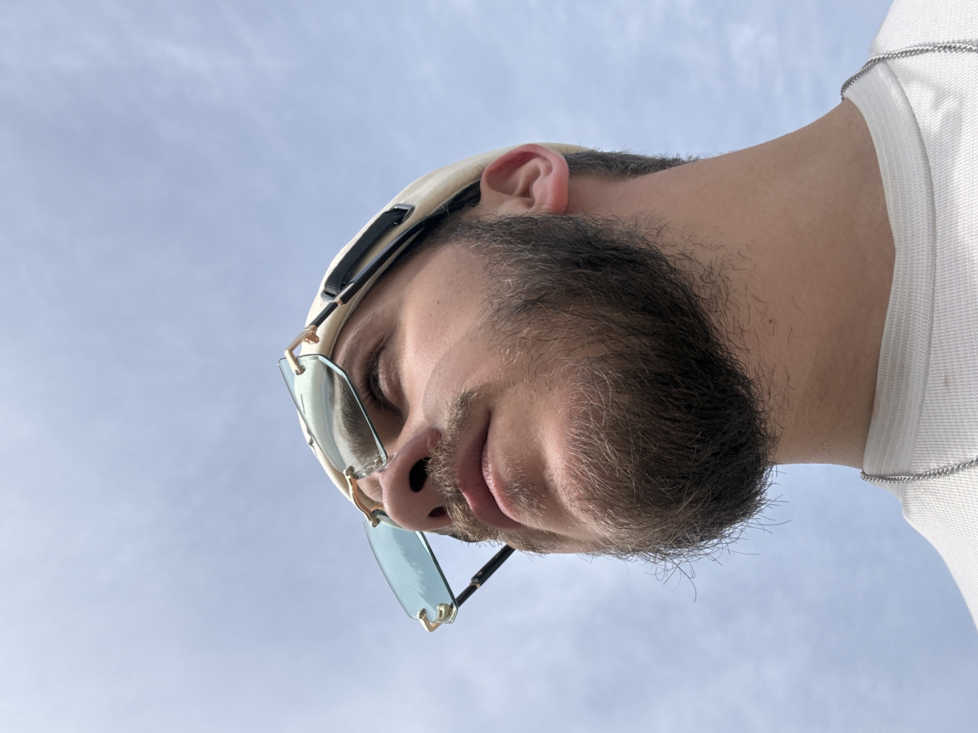 A man is viewed from below against a pale blue sky. He is wearing a beige baseball cap and light-tinted sunglasses. He has dark hair and a full, trimmed beard. He is wearing a white t-shirt with a silver chain detail at the collar. The image does not provide sufficient information to determine the location as Teteven, Bulgaria, or any other specific place.