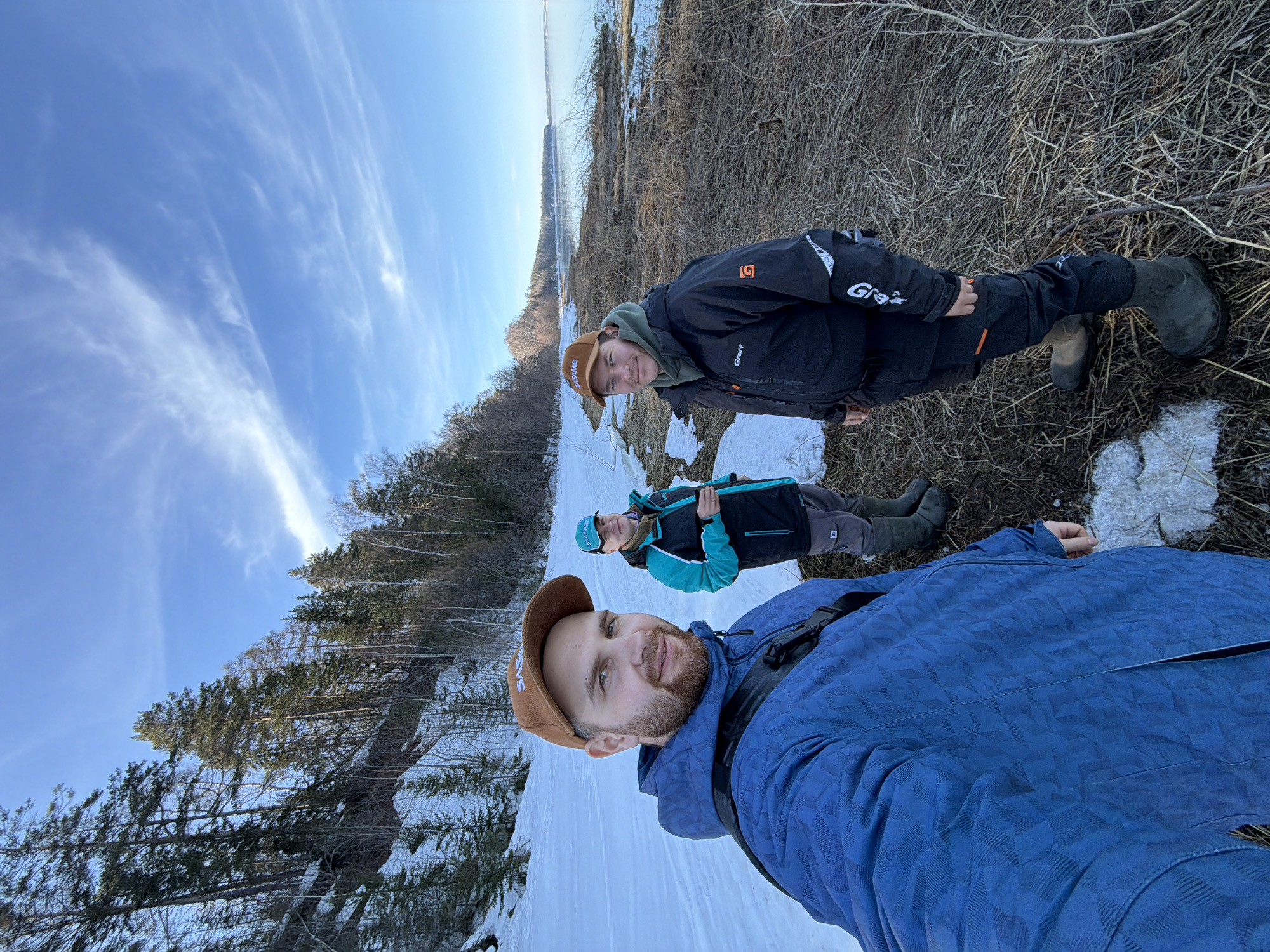 Three adult males are depicted in a selfie on a partially snow-covered shoreline next to a calm body of water. The location features a snow-dusted hillside on the left, densely covered with coniferous and bare deciduous trees. The ground consists of snow patches, exposed brown earth, and dried, light-brown vegetation extending to the water's edge. The water is calm, light blue-grey, and stretches to a distant treeline on the horizon. The sky above is bright blue with thin, wispy white clouds.
The individual holding the camera, positioned on the left, is a male with a light brown beard, wearing a blue patterned hooded jacket and a brown baseball cap with "SASAME" branding. A black strap from a cross-body bag is visible over his left shoulder. A second male, centrally located, wears a teal and black hooded jacket, a brown neck gaiter, grey trousers, and olive green rubber boots. He holds a dark-colored cup in his right hand. The third male, on the right, wears a black jacket with "Graff" branding and an orange "G" logo, an olive green hooded sweatshirt underneath, black trousers with an orange accent, and olive green rubber boots. He also wears a brown baseball cap with "SASAME" branding. All three subjects are dressed in heavy outdoor apparel suitable for cold weather. They are interacting by posing for the camera.