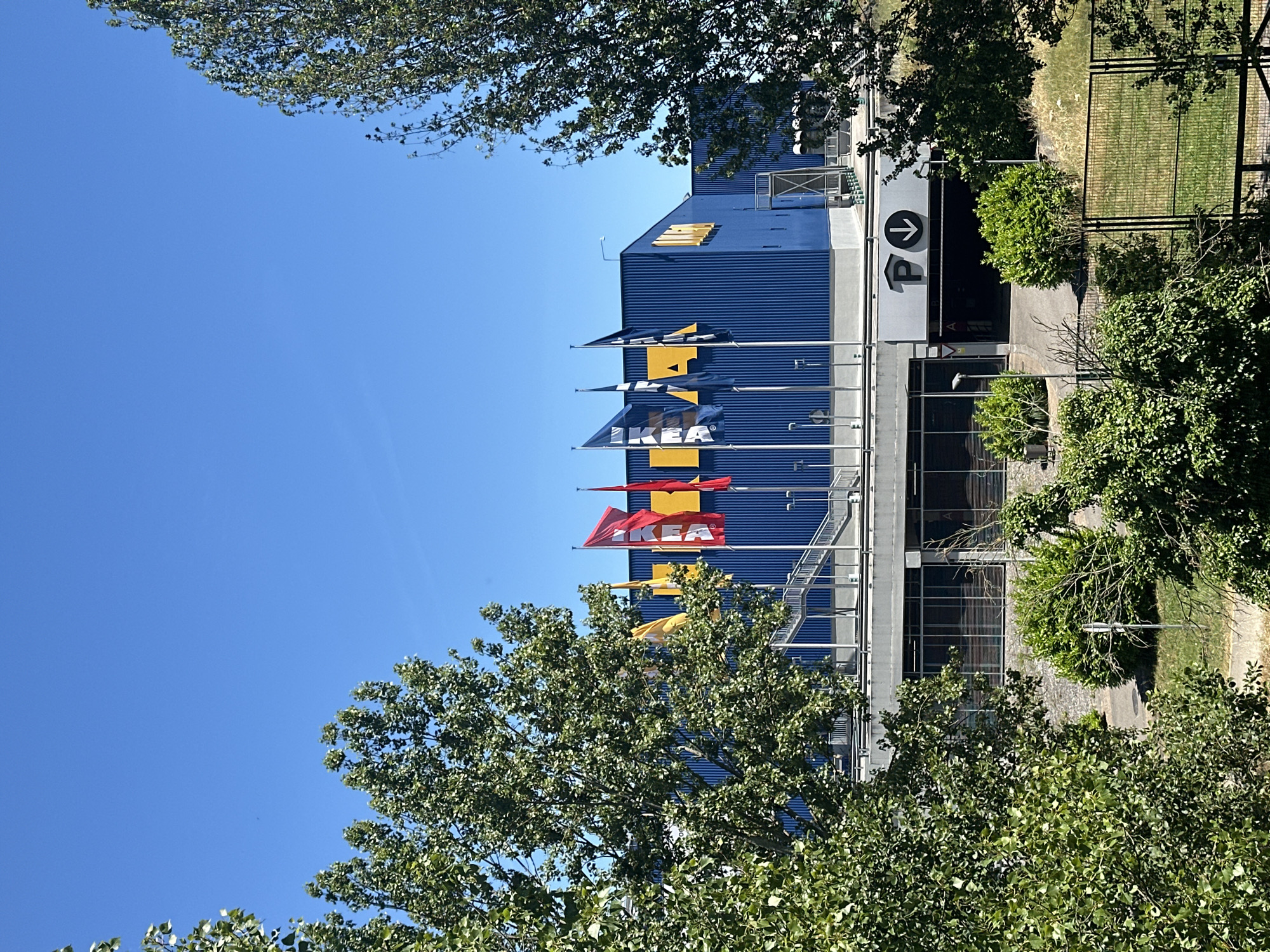 The image depicts the IKEA store located in the city of Oviedo, Spain. The facade of the store is predominantly blue, with large yellow IKEA logos visible on the upper portion. A series of flags, including red and yellow ones with the IKEA logo, are flying in front of the building. Stairs lead up to an entrance. Below the main store, there is a section with windows and an entrance to a parking garage, indicated by a "P" symbol with a downward arrow. The scene is framed by lush green trees on the left and right sides, and the sky above is clear and blue. The lighting suggests a bright, sunny day.