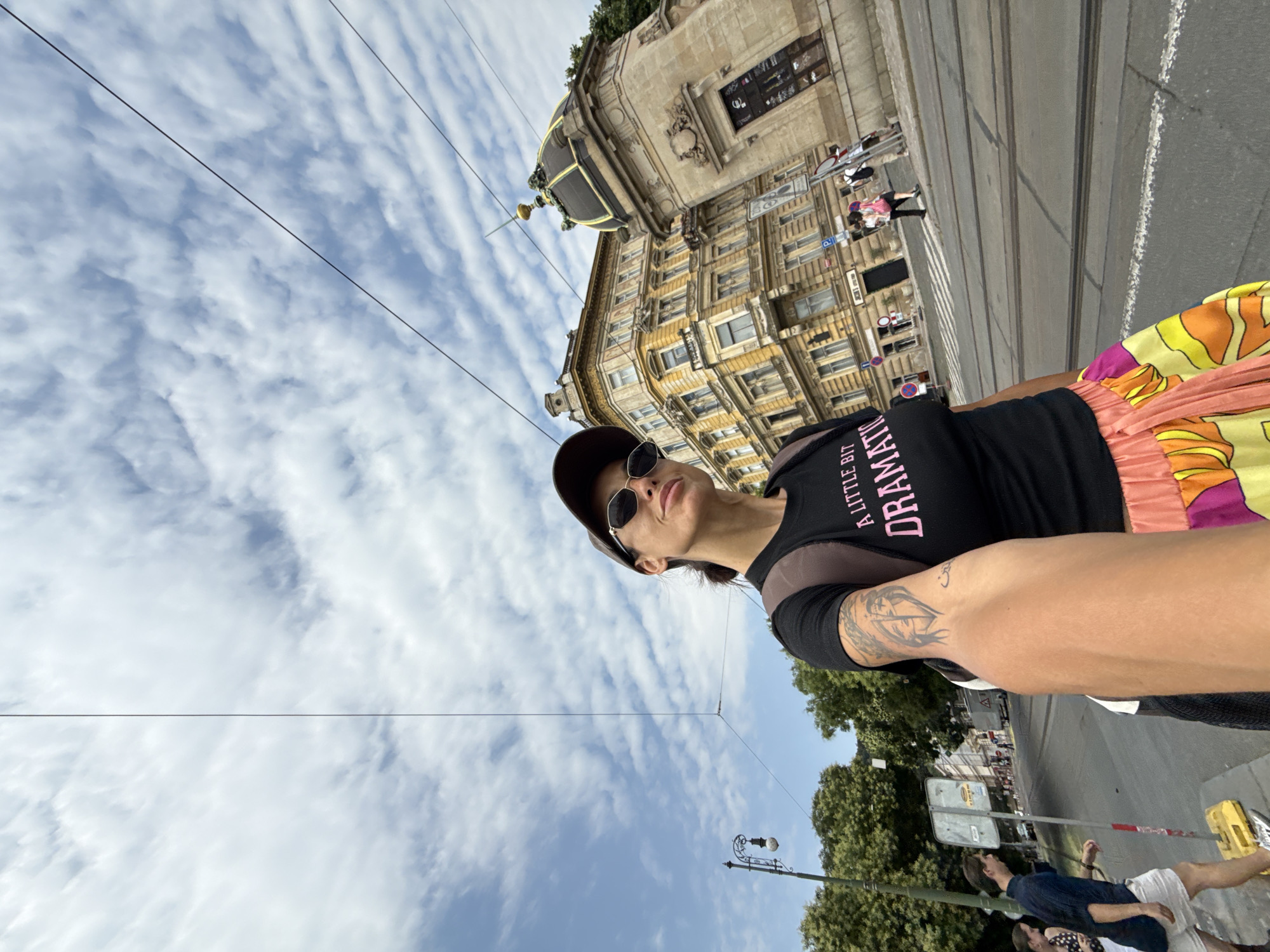 A woman, seen from the chest up and holding the camera with her right arm, takes a selfie on a sunny day in Prague, Czechia. She wears a black t-shirt with the words "A LITTLE BIT DRAMATIC" in pink lettering, a brown baseball cap, and sunglasses. She also has a backpack on her shoulders and tattoos on her left arm. Her skirt is colorful with a patterned design of orange, yellow, and pink.
The woman is standing on a street with tram tracks. In the background, there are several buildings, including a yellow building with many windows and a domed structure. Trees and a lamppost are also visible. There are other people in the background, walking on the sidewalk and crossing the street. The sky is blue with white clouds. The overall scene appears to be a busy urban environment.
