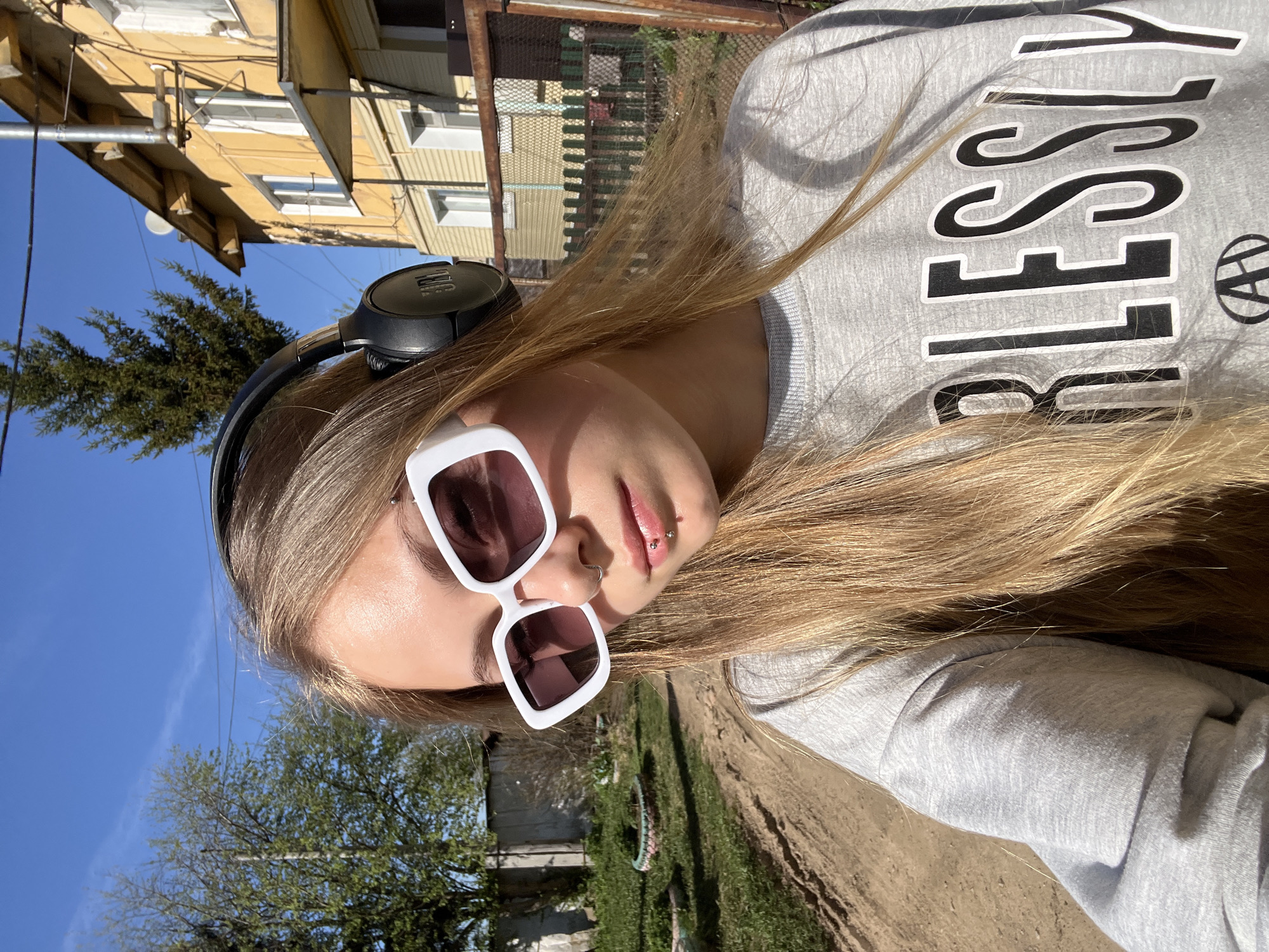 A young woman poses for a selfie outdoors in the city of Фоки, Russia. She is wearing large, square white sunglasses, a light gray sweatshirt with bold black lettering, and black headphones. She has long, blonde hair and facial piercings on her nose and lower lip. The background includes a yellow building with visible windows, a blue sky, trees, and a dirt road. A fence is also partially visible.