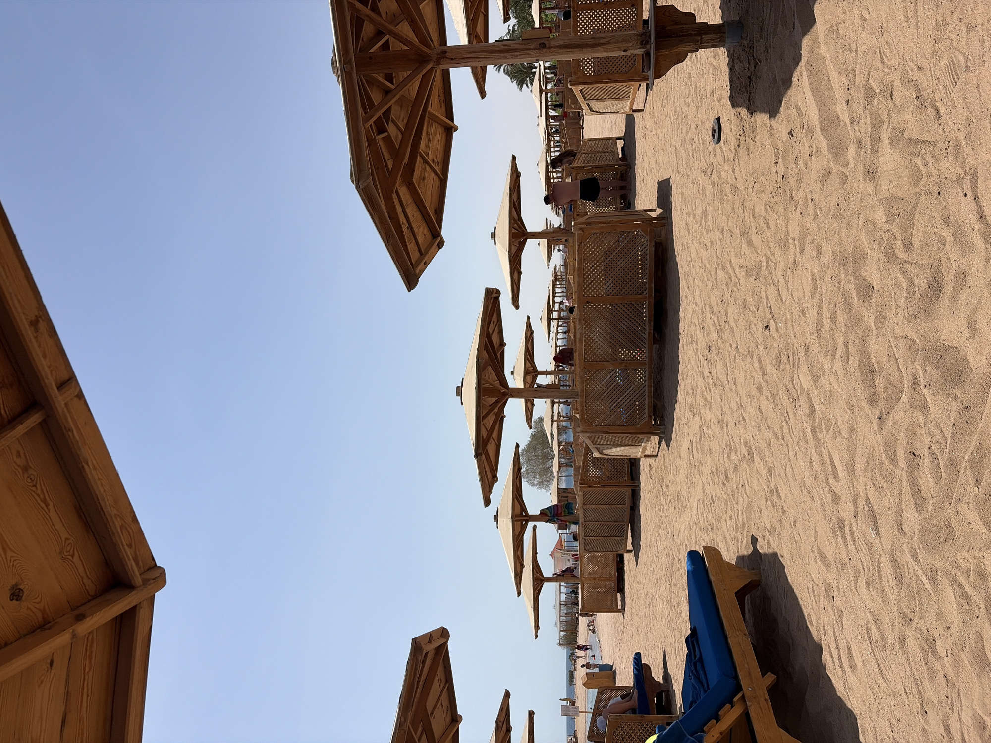 This image depicts a beach scene in Hurghada, Egypt, under a clear blue sky. The foreground is dominated by tan sand, with a blue lounge chair positioned on the left, casting a shadow. Several wooden umbrellas with thatched roofs are scattered across the beach, some with wooden privacy screens around them. In the background, the sea is visible, with a few people wading or standing in the shallow water. Other beachgoers are seen walking and sitting on lounge chairs. The overall atmosphere is one of a sunny day at a resort beach.
