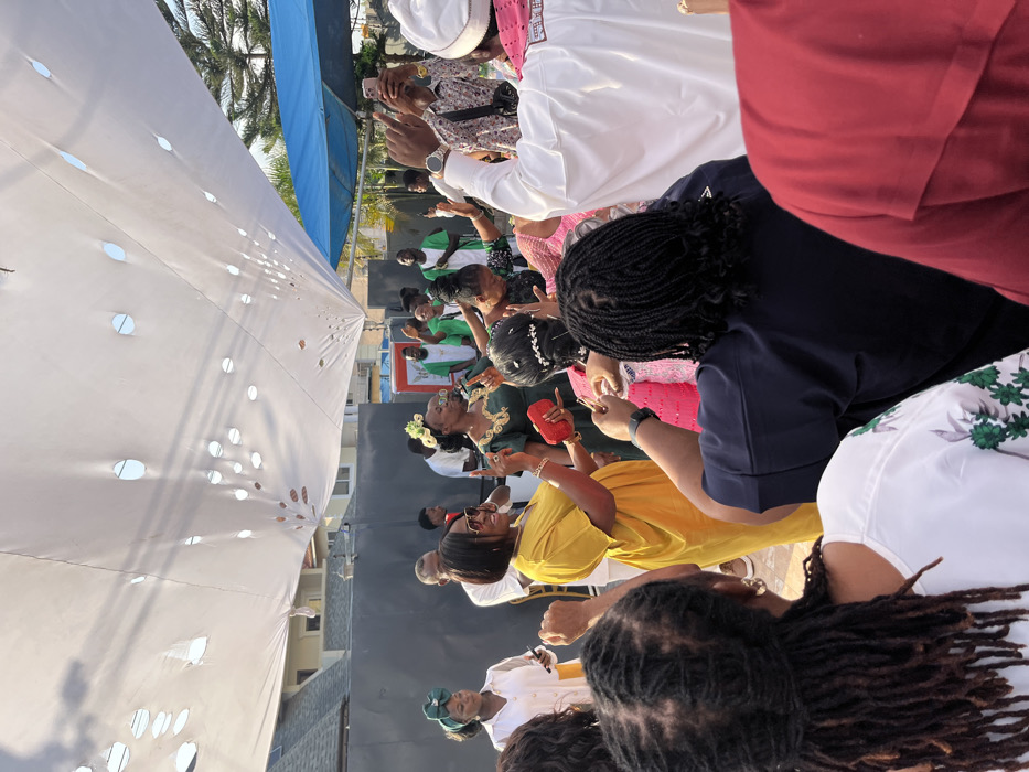 The image depicts an outdoor gathering in Unknown, Ghana, under a white fabric canopy featuring multiple circular perforations. Approximately 15-20 individuals, mostly adults, are visible, actively participating in what appears to be a celebratory or religious event.
In the midground, a woman in a yellow kaftan holds a red clutch bag and gestures with her right index finger, wearing sunglasses on her head. Adjacent to her, a woman in a dark green dress with gold embroidery and a floral headpiece stands with her eyes closed or cast downward, both hands raised in an expressive manner. Behind her, another woman in a pink garment also has raised hands.
To the left of the woman in yellow, a woman in a white shirt and green headwrap holds a smartphone. In the background, several men are present, some wearing white shirts with green sashes or trim. On the far right, a man in a white robe and white head covering holds a smartphone aloft to record the scene.
In the immediate foreground, with their backs to the viewer, are three individuals: one with dark dreadlocks wearing a white top with green floral patterns, one with dark braided hair in a dark blue top, and another partially visible in a red garment.
The ground consists of light-colored paving. The background shows dark wall-like structures, a light-colored building, a blue tarp or awning, and a palm tree, consistent with an urban or residential outdoor setting.