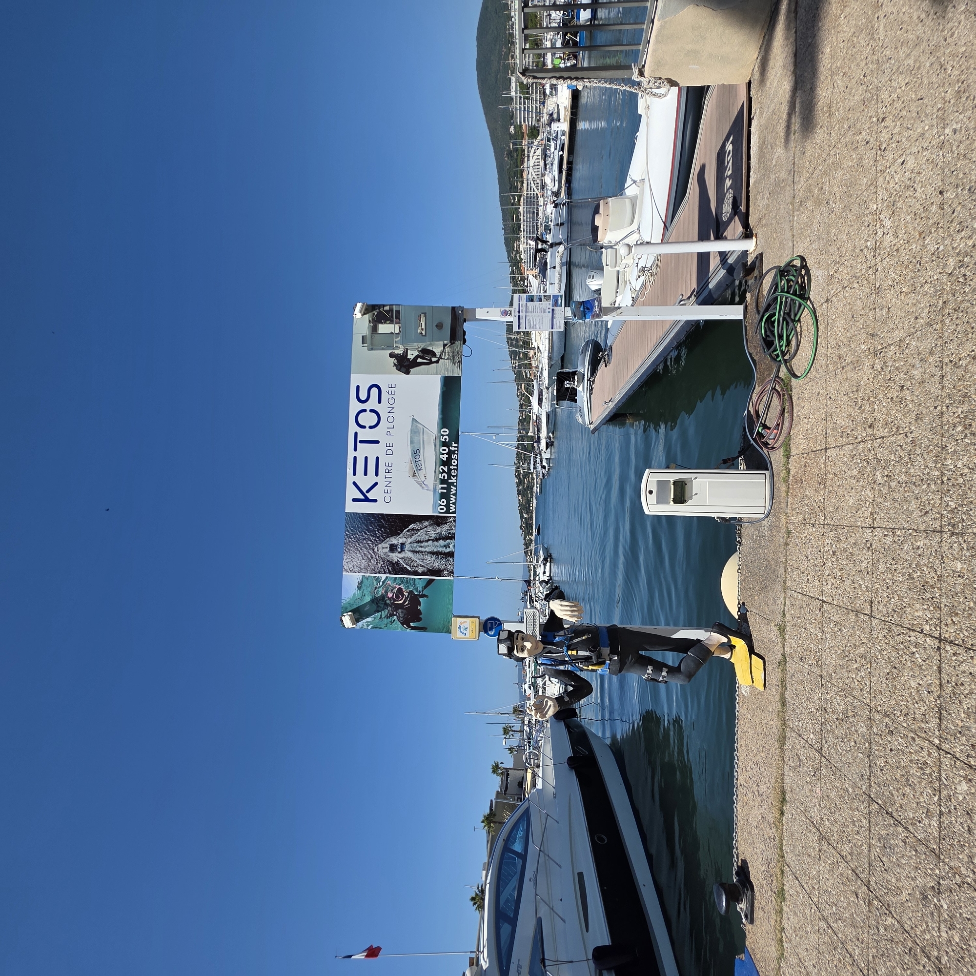 A diver statue is posed on a paved walkway by a harbor in Sainte-Maxime, France. The statue is wearing a black diving suit, yellow fins, and a blue diving vest, and is waving its right hand. Behind the statue, a large sign displays "KETOS CENTRE DE PLONGEE" along with images of divers and boats, and contact information. A French flag flies on a flagpole to the left. Several boats are docked in the harbor, including a white luxury yacht with blue-tinted windows and smaller boats on a wooden pier to the right. The sky is clear and blue, and a distant green hill is visible in the background.