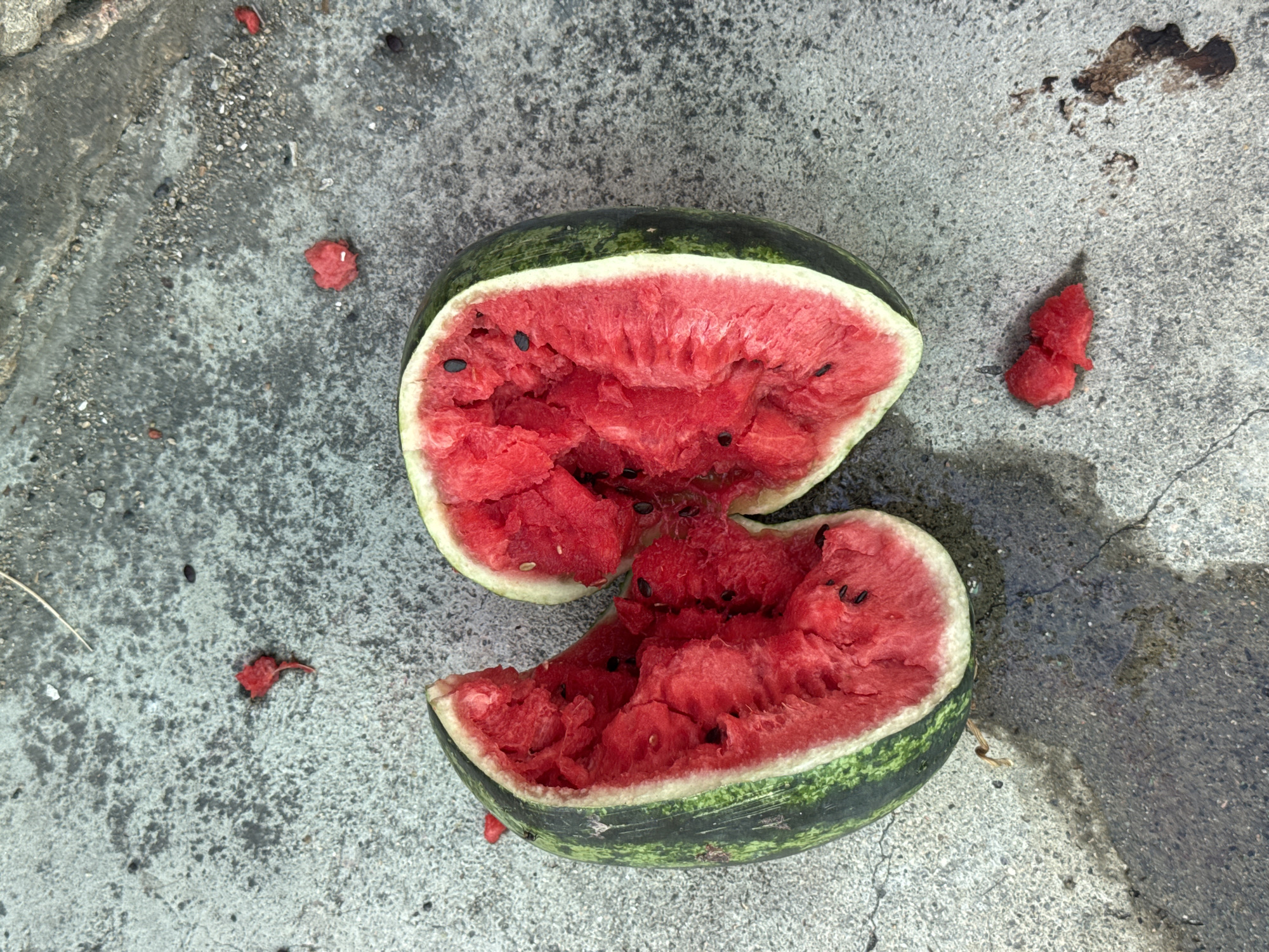 The image shows a split watermelon on a concrete surface. The watermelon is bright red and has black seeds. There are small pieces of watermelon flesh scattered around it, along with some dark stains on the concrete that appear to be watermelon juice. The concrete surface is rough and textured, with small pebbles and debris visible. The lighting suggests it is daytime, with shadows indicating sunlight. The scene appears to be outdoors, possibly in an urban setting given the concrete ground, but there is no clear indication of Nha Trang, Vietnam.