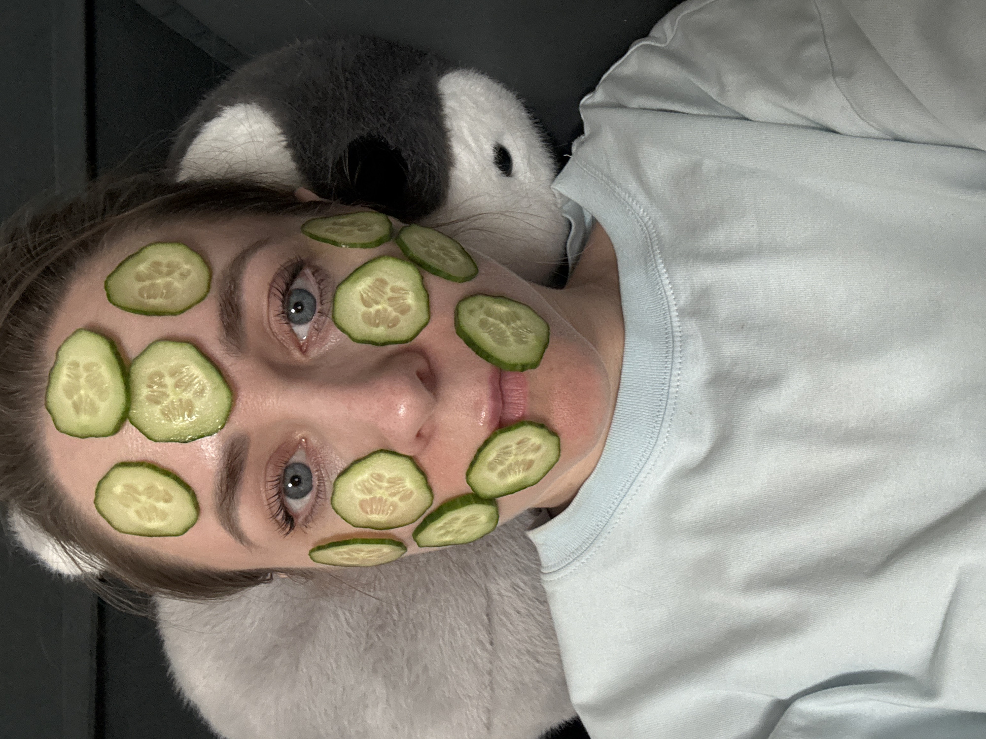 A woman is lying down with cucumber slices on her face. She has blue eyes, fair skin, and is wearing a light blue t-shirt. Her head rests on a soft, plush gray and white object, possibly a pillow or stuffed animal. The background includes a dark gray surface, potentially a couch. It is not possible to ascertain the location as the city of Chaikovsky, Russia, as the image contains no identifying information about a location.