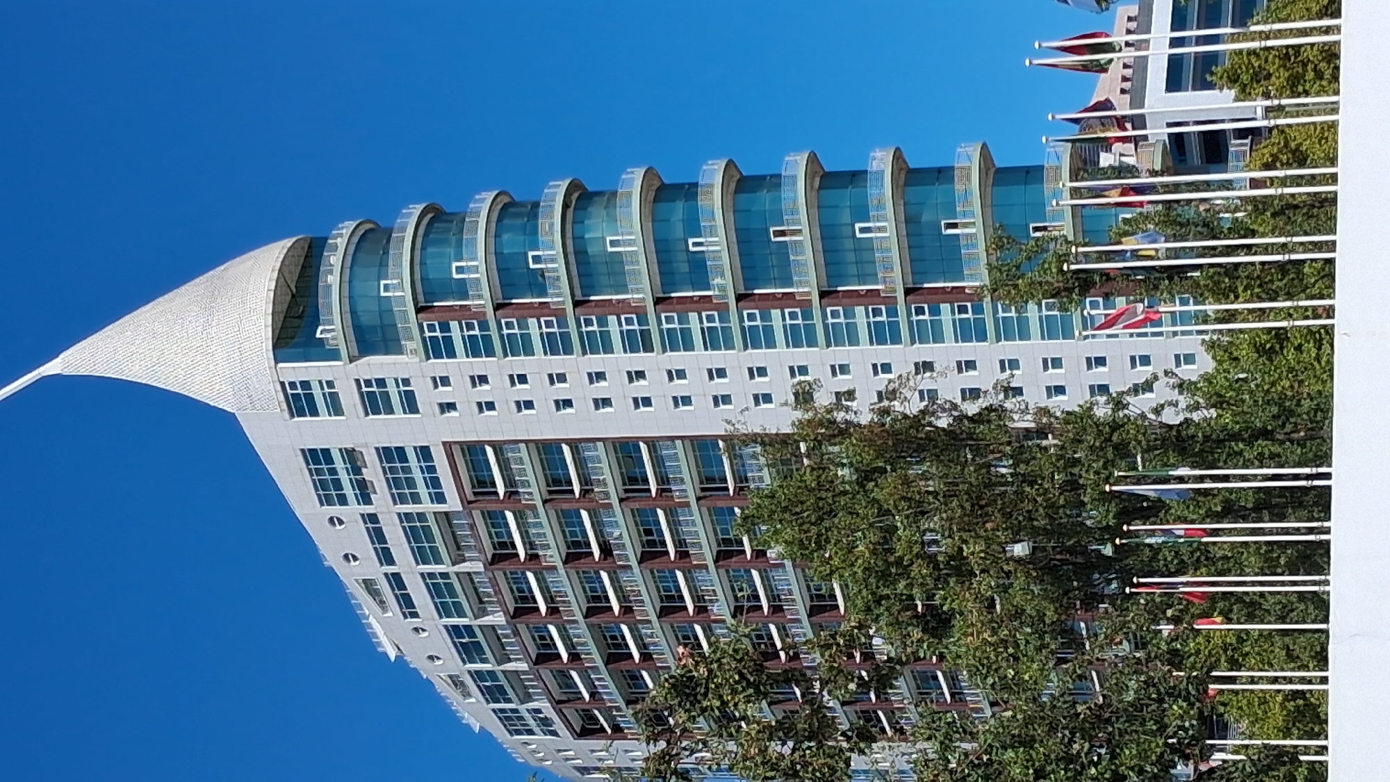 The image shows the iconic "Torre Vasco da Gama" (Vasco da Gama Tower), a skyscraper located in the Parque das Nações (Park of Nations) district of Lisbon, Portugal. The tower, with its distinctive sail-like silhouette, rises majestically against a clear blue sky. Its facade is a mix of light-colored panels and glass, with curved balconies that add a modern aesthetic.
At the base of the tower, a line of white flagpoles stands, each adorned with a different national flag, suggesting an international or diplomatic context. Lush green trees partially obscure the lower portion of the building, providing a contrast between nature and the modern architectural structure. The overall scene depicts a bright, sunny day in a developed urban area.