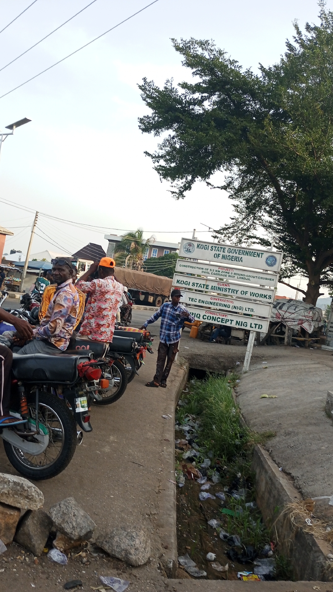 The image depicts a street scene in Adankolo, Nigeria. Several men are either sitting on or standing next to motorcycles parked along the edge of a road. A sign indicates that a road construction project is underway by the Kogi State Government of Nigeria. The project involves the construction of a 5.5 km zone and round-about/crusher dual carriage way in Lokoja. TEC Engineering (NIG) LTID is the contractor, and IQ Concept Nig LTID is the consultant. A truck is parked behind the motorcycles and a tree is in the background. A ditch filled with litter and grass runs along the side of the road.