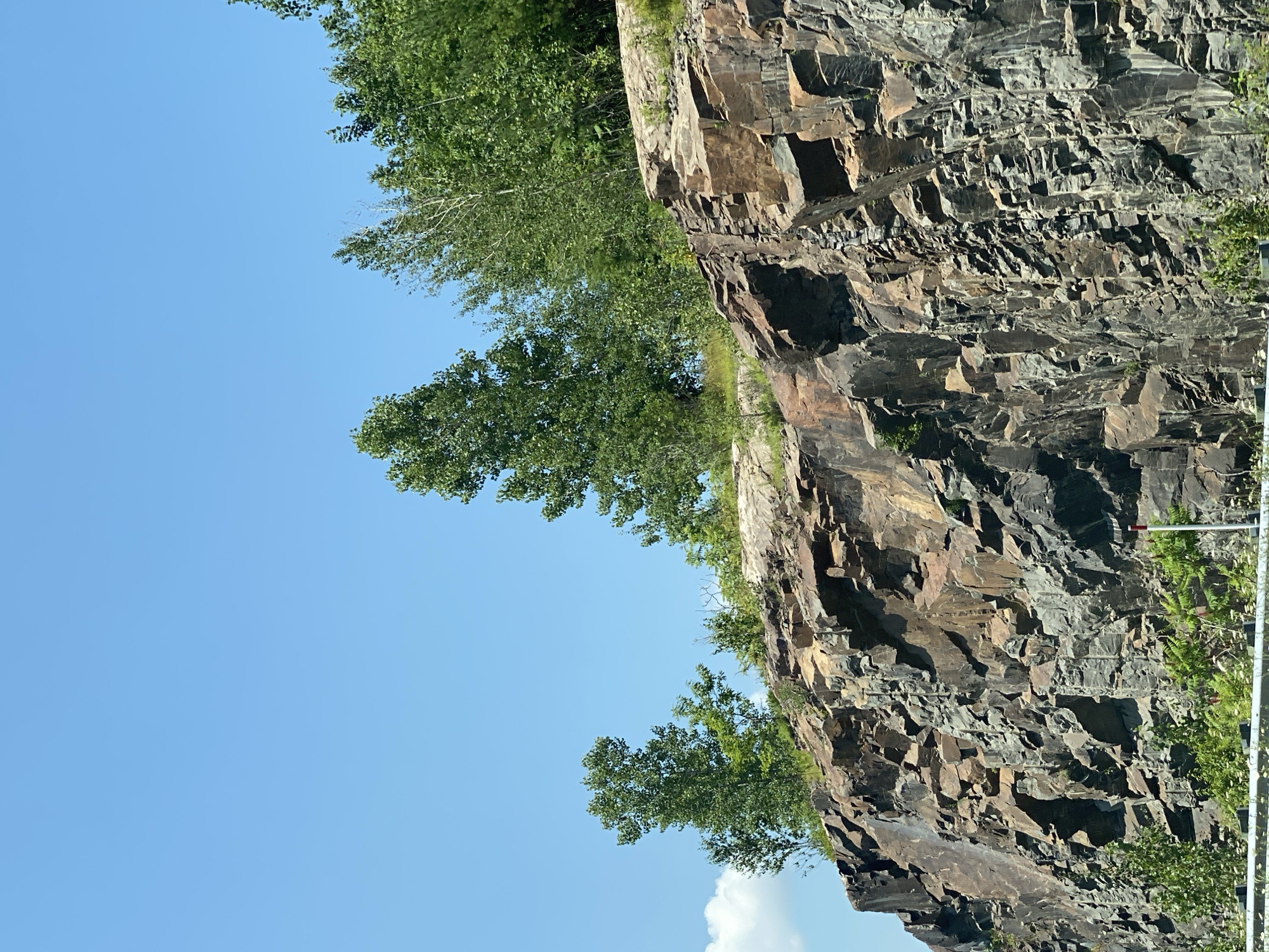 The image depicts a large, rugged rock face under a clear blue sky. The rock formation is primarily composed of dark, angular stones with some lighter, reddish-brown sections visible, suggesting varied mineral content or weathering. Vegetation, including trees and smaller green plants, grows on top of and at the base of the rock face. The trees on top are lush and green, with various heights and leaf densities. At the bottom of the image, a white barrier or railing is visible, along with some greenery. A white and red striped pole, possibly a marker, is also present near the bottom right. The overall scene appears to be a natural landscape, likely a roadside cliff, within the municipality of Grenville-sur-la-Rouge, Canada, given the context provided. There are no people or animals visible in the image.
