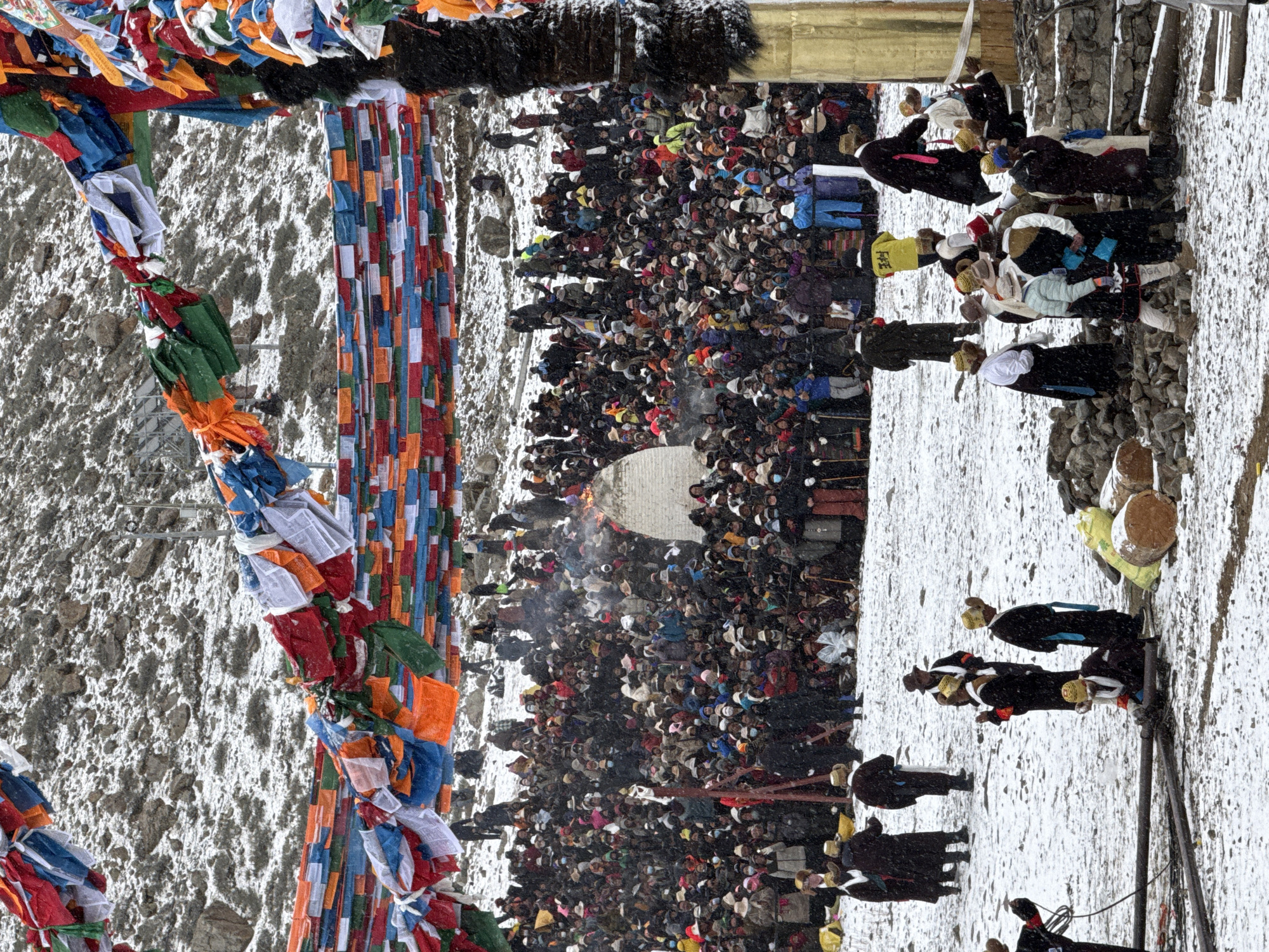The image depicts a large crowd of people gathered in a snowy environment. Prayer flags are prominently displayed on the left side of the image, adding color to the scene. The crowd appears to be focused on a central area where smoke or steam is visible, possibly indicating a ritual or event involving fire. Individuals in the foreground wear distinct traditional clothing, including robes and headdresses. The ground is covered in snow, and the background reveals a rocky hillside. The image is rotated counterclockwise. The location is suggested to be in Parga town, China.