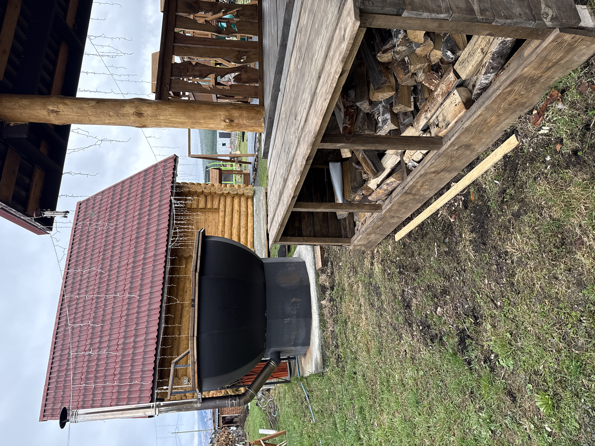 This image, captured in Tyulyuk, Russia, showcases a rustic outdoor setting dominated by functional structures under an overcast sky. In the foreground and left, a large, dark-colored outdoor oven or smoker, possibly made of metal, is prominently featured, resting on a concrete base. A shiny silver metal chimney pipe extends upwards from its side.
Above the oven, a steeply sloped red corrugated metal roof is visible, adorned with unlit string lights, suggesting a festive or decorative touch. To the right of the oven, a sturdy wooden structure, likely a woodshed or an extension of a covered porch, is filled with neatly stacked firewood. A prominent, rough-hewn wooden beam spans horizontally across the upper middle of the frame, adding to the rural aesthetic.
In the mid-ground, a log cabin-style building can be seen, further emphasizing the natural, perhaps traditional, architectural style of the location. The ground is a mix of sparse green grass and darker patches of earth, indicating a natural, unmanicured landscape. The sky is uniformly overcast and grey, suggesting a cloudy day without direct sunlight. There are no people visible, and no specific activity is taking place, presenting a still snapshot of an outdoor area prepared for use. No text is visible in the image.