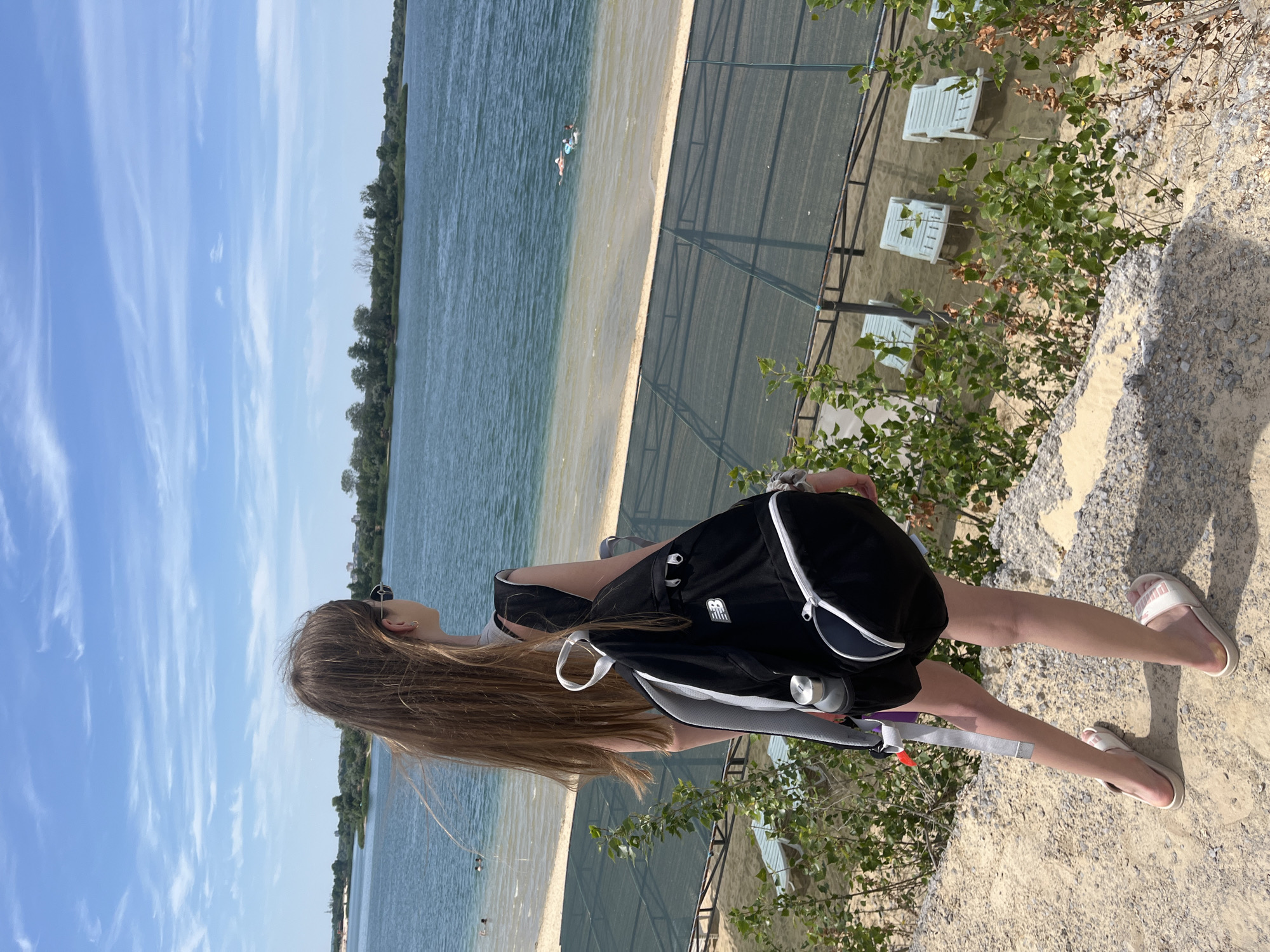 The image shows a young woman with long, light brown hair standing on a sandy embankment overlooking a body of water. She is wearing a black backpack with white trim, shorts, and light-colored slides. The backpack has a white logo with blue lettering. The woman is looking out towards the water, her head turned to the side.
The scene is a beach area with a sandy shore and clear blue water. There are several people in the water in the distance, appearing to be swimming or relaxing. Behind the beach, there is a fenced area with white lounge chairs and some greenery. The sky is blue with scattered white clouds, suggesting a sunny day. The overall impression is of a leisurely summer day at a beach resort.
There is no visible text in the image, other than the logo on the backpack which is partially obscured. The image appears to have been taken in daylight, likely during the afternoon due to the bright sunlight casting shadows. The location appears to be a recreational area, possibly a lake or a large quarry repurposed for swimming, given the clear water and sandy shore. The woman's pose and the relaxed atmosphere suggest a calm and enjoyable outing.