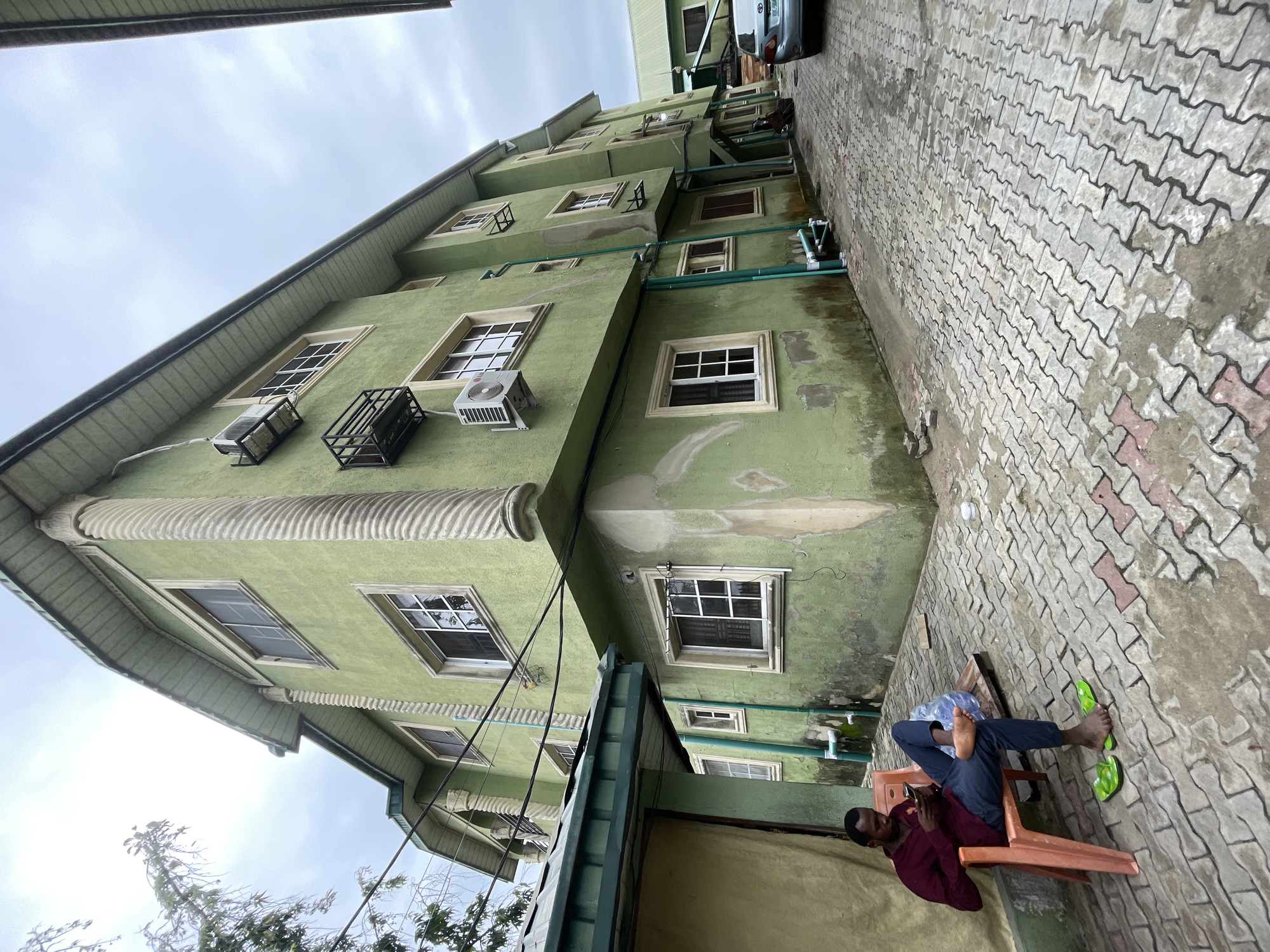An outdoor scene shows a four-story green building with white-framed windows, several with visible air conditioning units. Green piping runs along the exterior. To the right of the building, the ground is covered with interlocking pavement bricks. A man wearing a burgundy shirt, dark pants, and green flip-flops sits in an orange chair on the pavement. He is looking at a phone in his hand. The sky is overcast. The scene is in Ado, Nigeria.