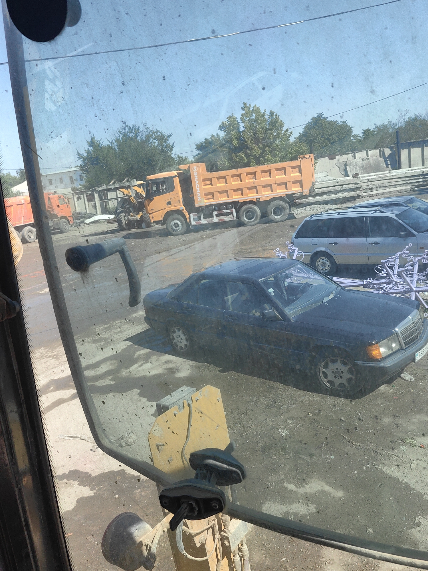 The image, taken from inside a construction vehicle cab, shows an outdoor scene with several vehicles and construction materials. In the background, an orange dump truck is parked next to a yellow excavator. In the foreground, a dark-colored sedan, possibly a Mercedes-Benz, is parked on a dirt and gravel surface. A silver station wagon is parked behind the sedan. The area appears to be a construction site or an industrial yard, with piles of concrete slabs visible in the background. The sky is blue and clear, suggesting daylight. The image may have been taken in the city of Shymkent, Kazakhstan, given the context.