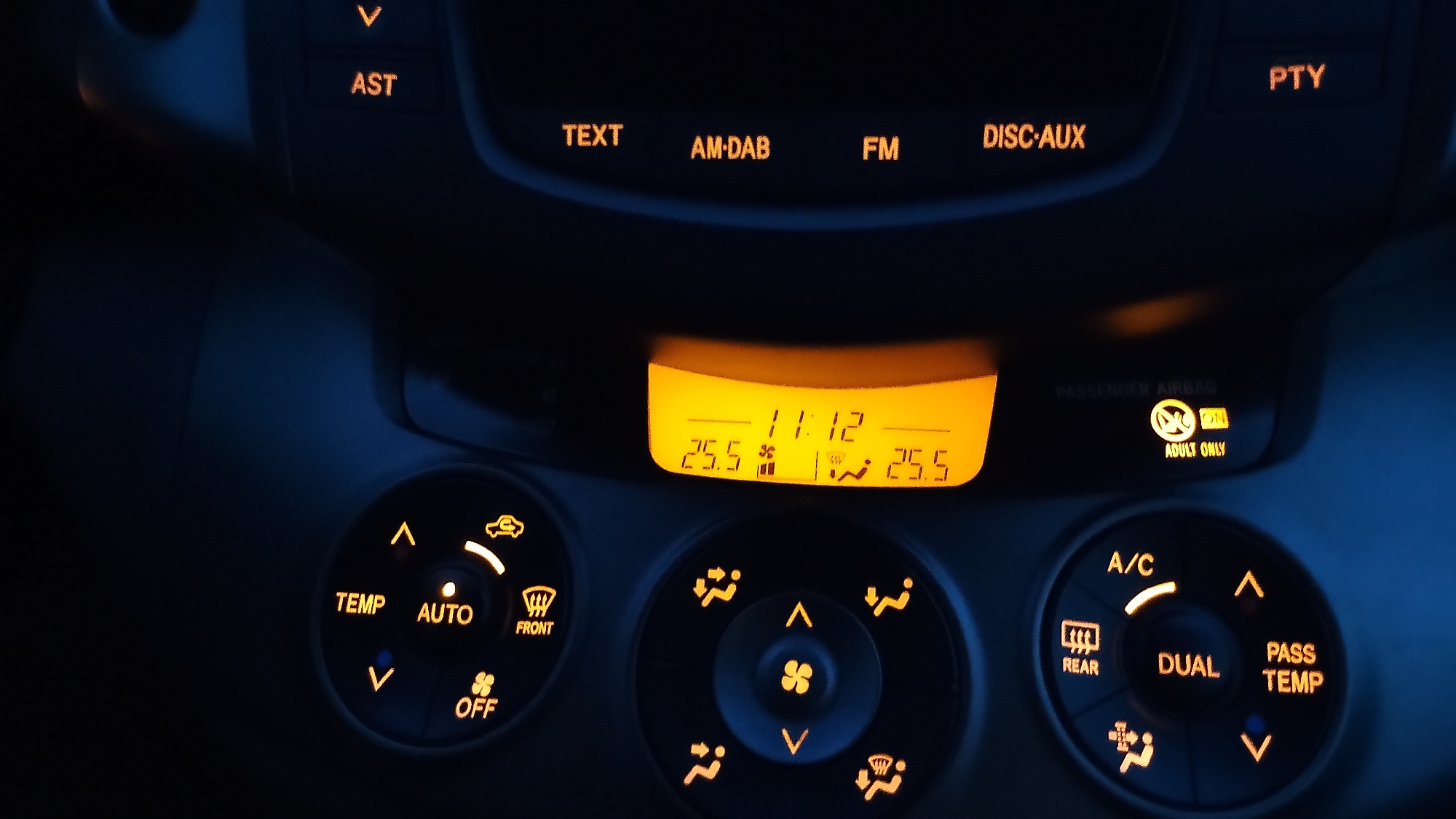 The image shows the interior of a car at night, focusing on the dashboard. The dashboard features an orange-lit digital display showing the time as "11:12" and two temperature readings of "25.5". Below the display are various controls for the climate system, including buttons labeled "TEMP", "AUTO", "OFF", "FRONT", "A/C", "REAR", "DUAL", and "PASS TEMP". There are also illuminated symbols indicating fan speed and air direction. Additionally, some buttons for the car's audio system are visible at the top of the dashboard, labeled "AST", "TEXT", "AM-DAB", "FM", "DISC-AUX", and "PTY". A small orange light illuminates an "ADULT ONLY" indicator. The car appears to be in a dark environment, suggesting it is parked or driving at night. No specific details about the city of Tazha, Bulgaria, can be inferred from this image.