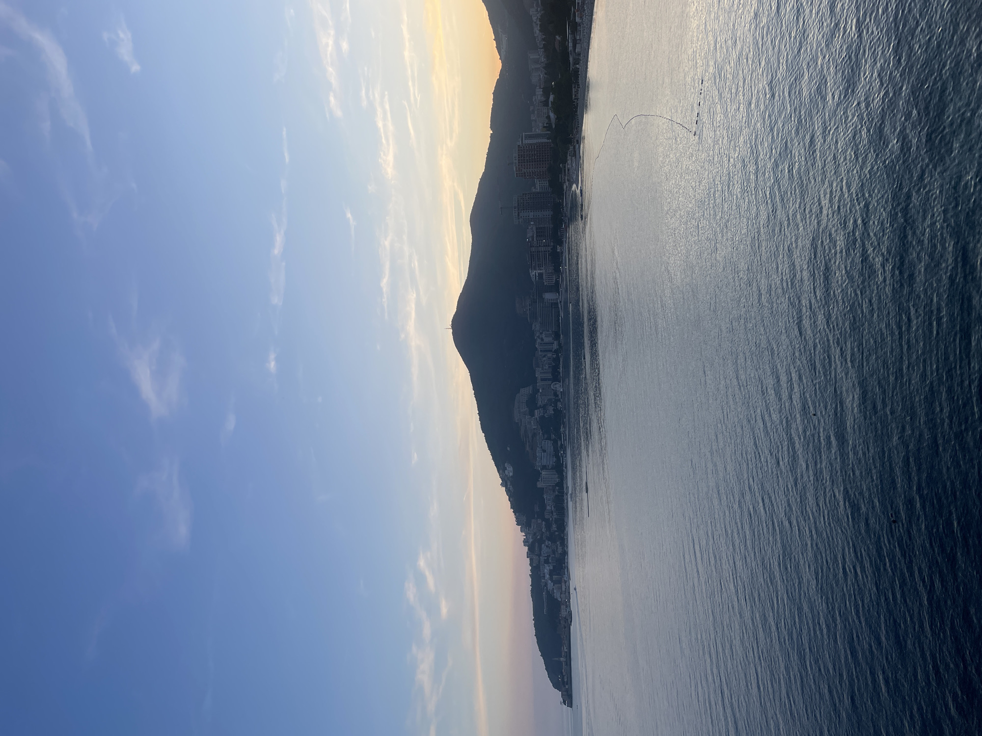 The image displays a panoramic view of a coastal landscape during either sunrise or sunset. The upper half is dominated by a clear blue sky, which lightens to a soft, golden hue towards the right horizon. Scattered wispy white clouds are visible across the sky.
A prominent range of mountains forms a dark silhouette in the midground, outlining the coastline. At the foot of these mountains, a densely built city extends, with numerous high-rise buildings and structures also appearing in silhouette against the brighter horizon.
The lower half of the image features a vast body of water, likely an ocean or bay, characterized by a rippled, textured surface reflecting the ambient light. A distinct, curved line of small, dark objects, possibly buoys or a series of small boats, is present on the water in the middle-right section.
The scene exudes a sense of tranquility, highlighting the interplay between the natural mountainous terrain, urban development, and the sea under the warm, transitional light of daybreak or dusk. No specific activities or visible text are apparent in the image.