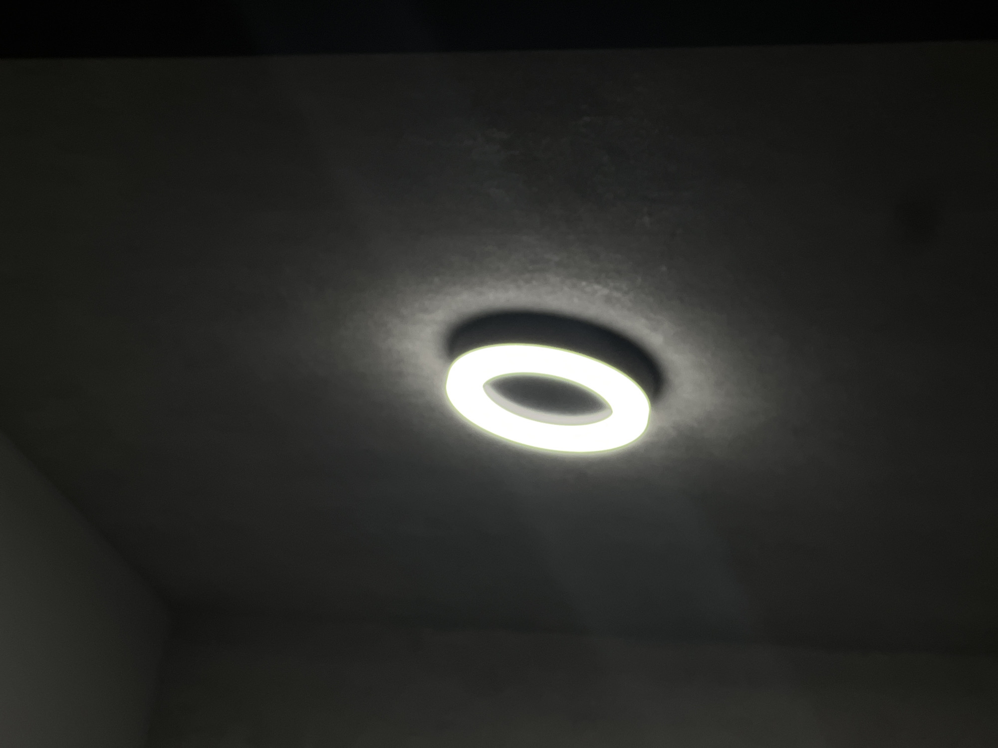 A glowing, white, illuminated letter "O" is affixed to a dark, textured wall, possibly in an indoor or outdoor urban setting. The "O" appears to be a sign or part of a larger display. The lighting suggests it is nighttime or a dimly lit area. The overall scene is monochromatic, with the bright glow of the "O" contrasting with the dark background. The location is identified as Sheptytskyi, Ukraine.
