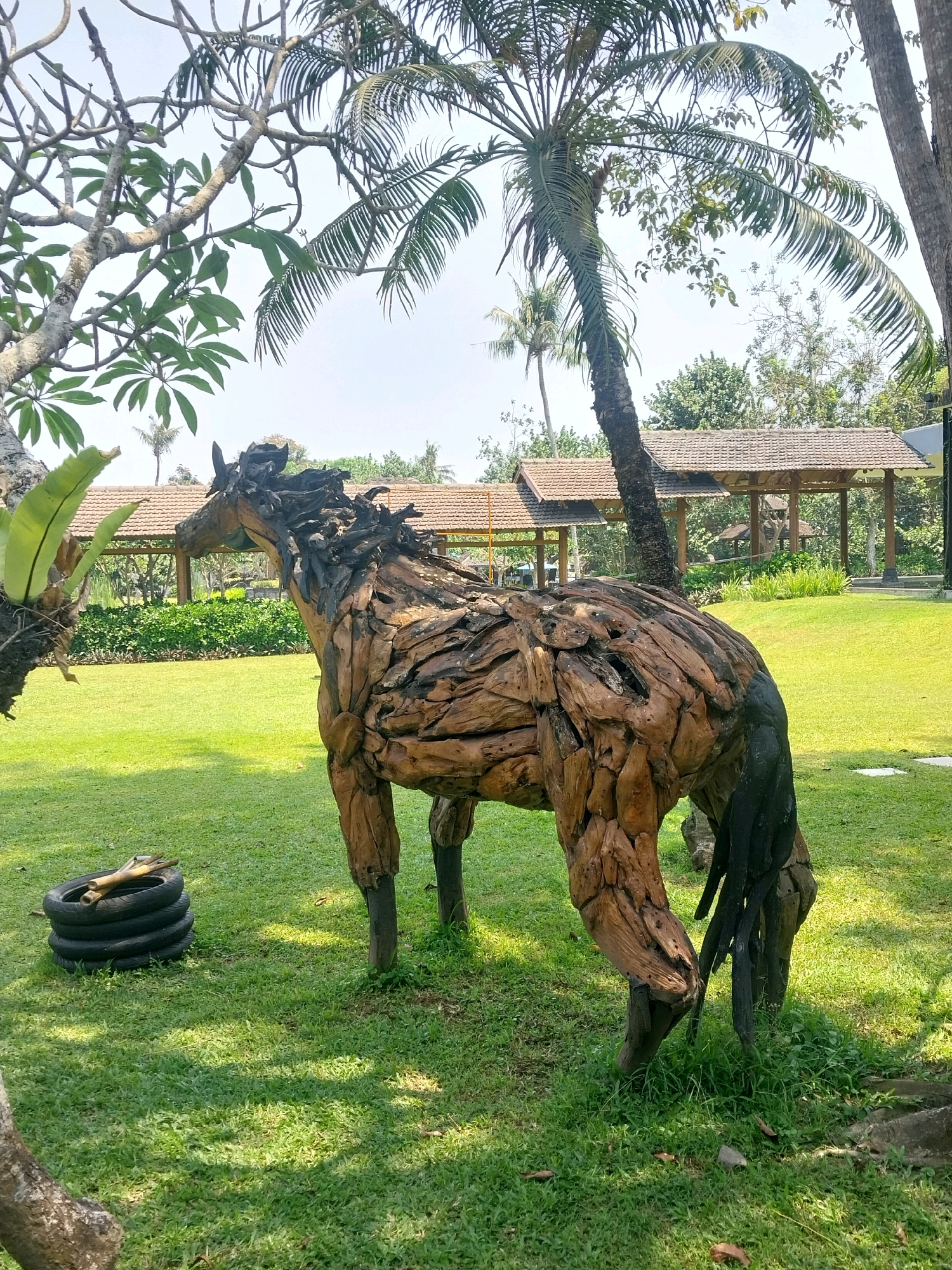 This is a medium shot, eye-level, outdoor photograph taken in Ngaglik, Indonesia. The subject is a large, rustic sculpture of a horse, meticulously crafted from reclaimed wood, positioned on a vibrant green lawn. The sculpture has a textured, natural appearance, with individual wooden pieces forming its body, mane, and tail. One of the horse's hind legs is partially painted black. To the left of the horse, a stack of black tires is visible on the grass.
In the background, a picturesque scene unfolds with lush greenery and traditional Indonesian architecture. Several palm trees are prominent in the frame, with their fronds gently swaying. A series of open-air gazebos or pavilions with tiled roofs are visible, connected by walkways. The overall environment suggests a well-maintained garden or resort area, bathed in natural daylight. The sky above is clear and bright, indicating fair weather. No people are present in the image.