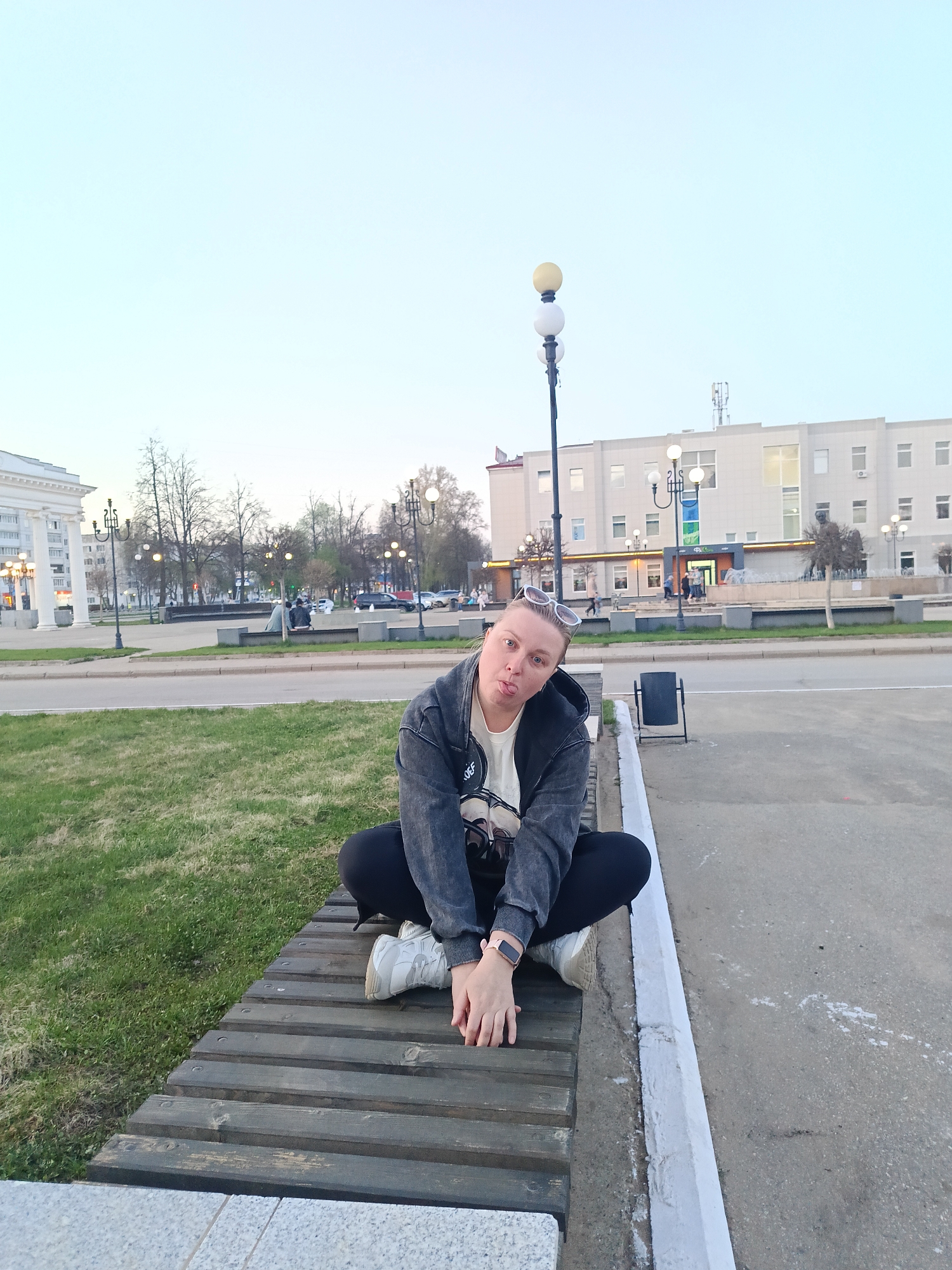 A woman is sitting cross-legged on a wooden bench in Chaikovsky, Russia. She is wearing black pants, white sneakers, a white t-shirt, and a grey jacket. She has sunglasses perched on her head and is sticking her tongue out.
The bench is situated in a paved public area with grass nearby. In the background, there is a multi-story light-colored building with several windows and a fountain in front. A street with cars and pedestrians is visible between the bench and the building. Streetlights are present, and the sky is overcast. In the left background, classical architectural features with pillars are visible. A trash can sits adjacent to the bench.