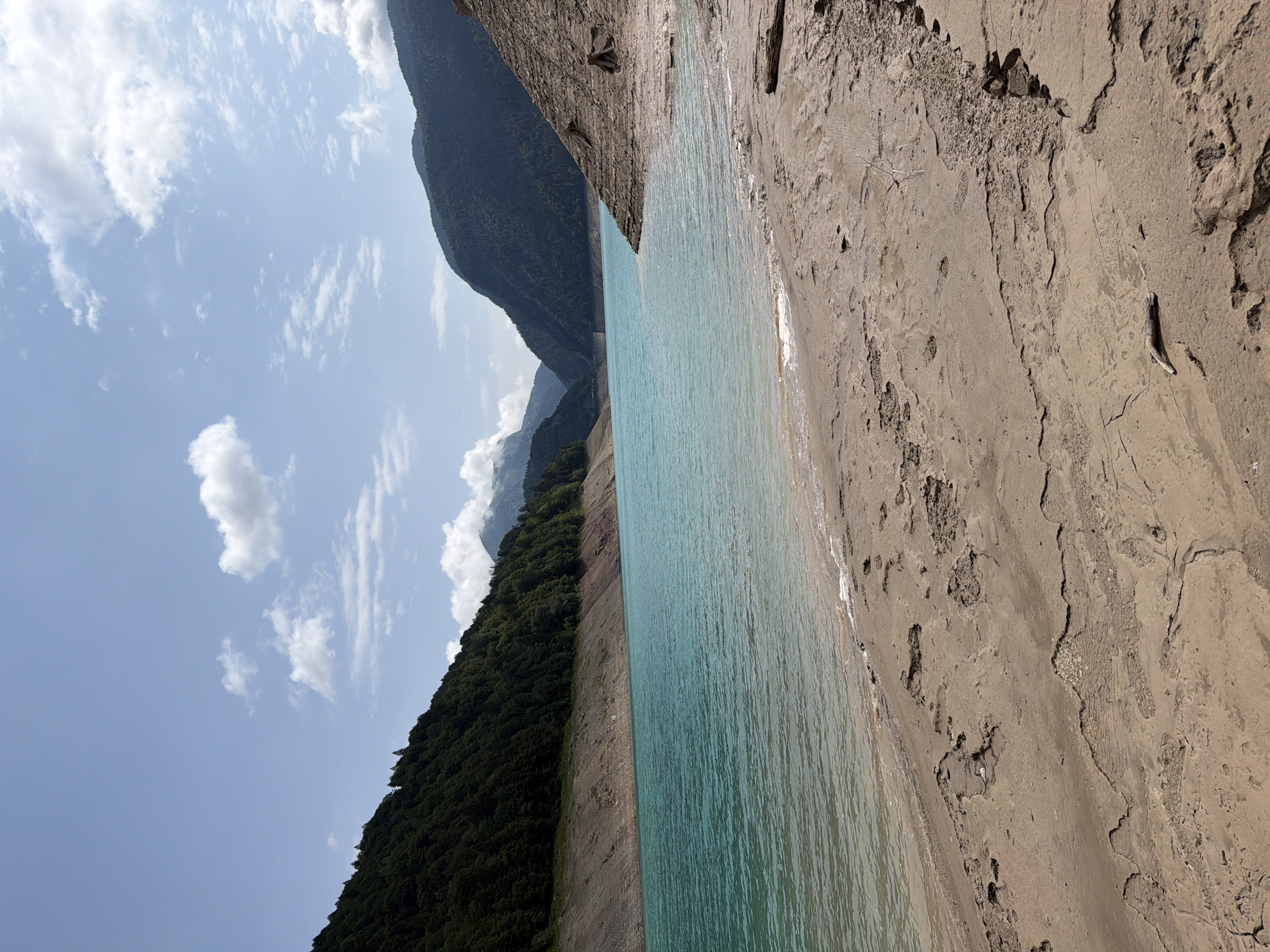 A wide shot captures a serene lake in Sauris di Sopra, Italy, under a partly cloudy sky. The turquoise water stretches across the frame, meeting a sandy shore in the foreground. The shore is marked by various impressions, some appearing like dried mud cracks, others like footprints. To the left, a densely forested mountain slope rises steeply from the water's edge. On the right, a rocky, barren slope dominates the foreground, with a few dead tree stumps emerging from the soil. In the distance, more mountains, also covered in trees, form a backdrop, with a visible dam structure in the far distance. The scene is devoid of any people or animals.