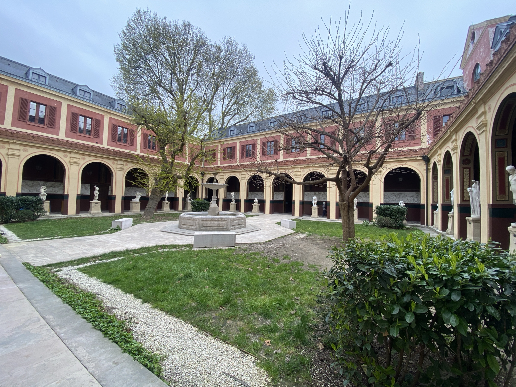 The image depicts the courtyard of the Musée Carnavalet, located in Paris, France. The courtyard features a central fountain surrounded by grass and stone pathways. Buildings enclose the courtyard, displaying arched openings with statues positioned within them. The buildings have a red and beige exterior, with dormer windows along the roofline. Two bare trees are in the courtyard, one to the right of the fountain and one behind it. Bushes and small plants are visible in the foreground and along the pathways. The sky is overcast. No people are explicitly visible in the image.