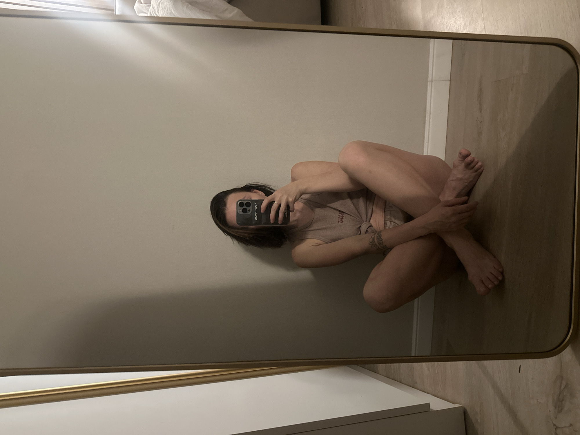 A woman is seen in a mirror. She is sitting on the floor with her legs crossed and arms around her knees. She is holding a phone in her hand, obscuring her face, and taking a picture of herself in the mirror. The mirror is framed in gold and the wall behind the woman is beige. The floor is made of wood planks. The photo appears to be taken indoors. This scene is not in Votkinsk, Russia.