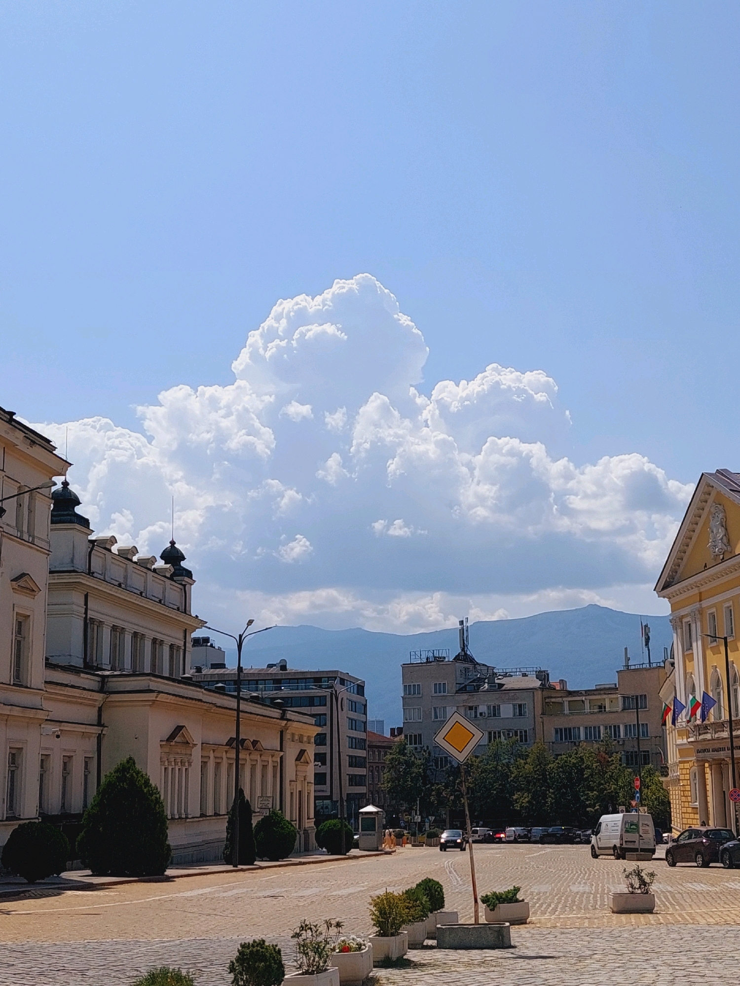 This is a photograph of a city square in Sofia, Bulgaria, likely located near the National Assembly building. In the foreground, a cobblestone street is visible, bordered by planters with manicured bushes. Several buildings line the street. On the left is a light-colored building with classical architectural elements, including columns and decorative cornices. To the right, a prominent yellow building with an ornate facade and flags flying from its balconies stands out. In the middle ground, there are more modern buildings, trees, and a traffic intersection with cars, including a white van. A diamond-shaped traffic sign is positioned near the intersection. In the background, a range of blue mountains forms the horizon. Above the cityscape, a dramatic cloud formation dominates the bright blue sky. The scene is captured during daylight, suggesting fair weather.