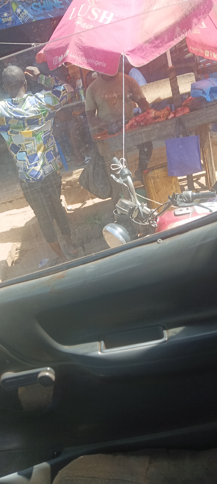 The image shows a scene in Kuru Dungus, Nigeria, viewed from inside a car. Through the car window, a street-side meat market is visible, shaded by a pink umbrella with the inscription "LUSH Hair" and the handle "Lushhairnigeria". Two men are present; one is wearing a patterned shirt and the other a plain green shirt. The man in the green shirt is behind a table with raw meat on it. A black motorcycle is parked in front of the table. The background also includes other commercial signs and street-side objects. The interior of the car with a gray door panel and door handle is visible in the foreground.