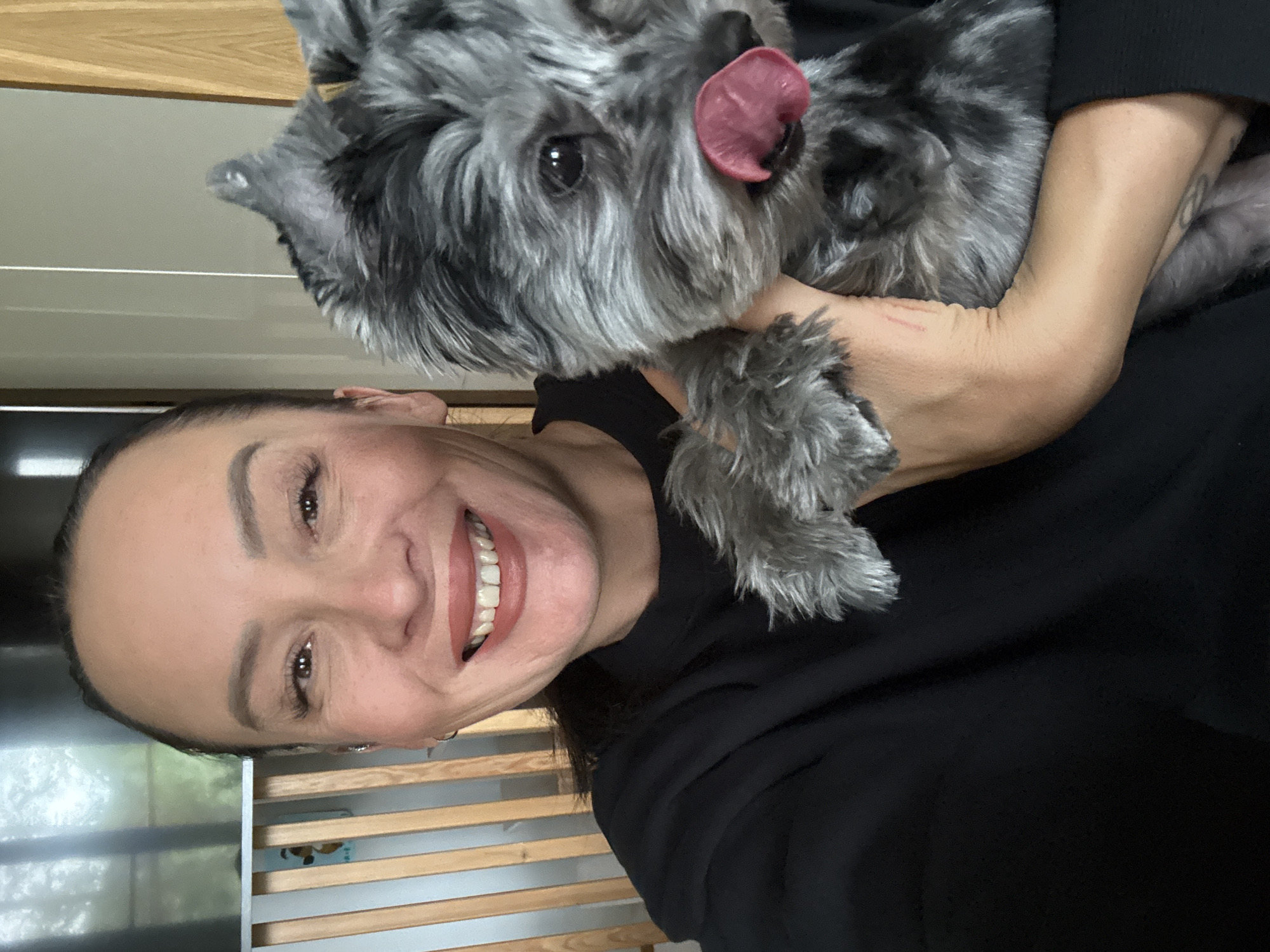 A woman with dark hair pulled back and wearing a black sweater holds a small, grey dog with its tongue sticking out. The woman is smiling broadly and looking at the camera. The dog is held in her arms, facing towards the camera, with its head tilted slightly to the right. The background consists of wooden slatted panels and a blurred interior. The image appears to be a selfie taken indoors. There is no discernible location in the city of Chaikovsky, Russia, in this image.