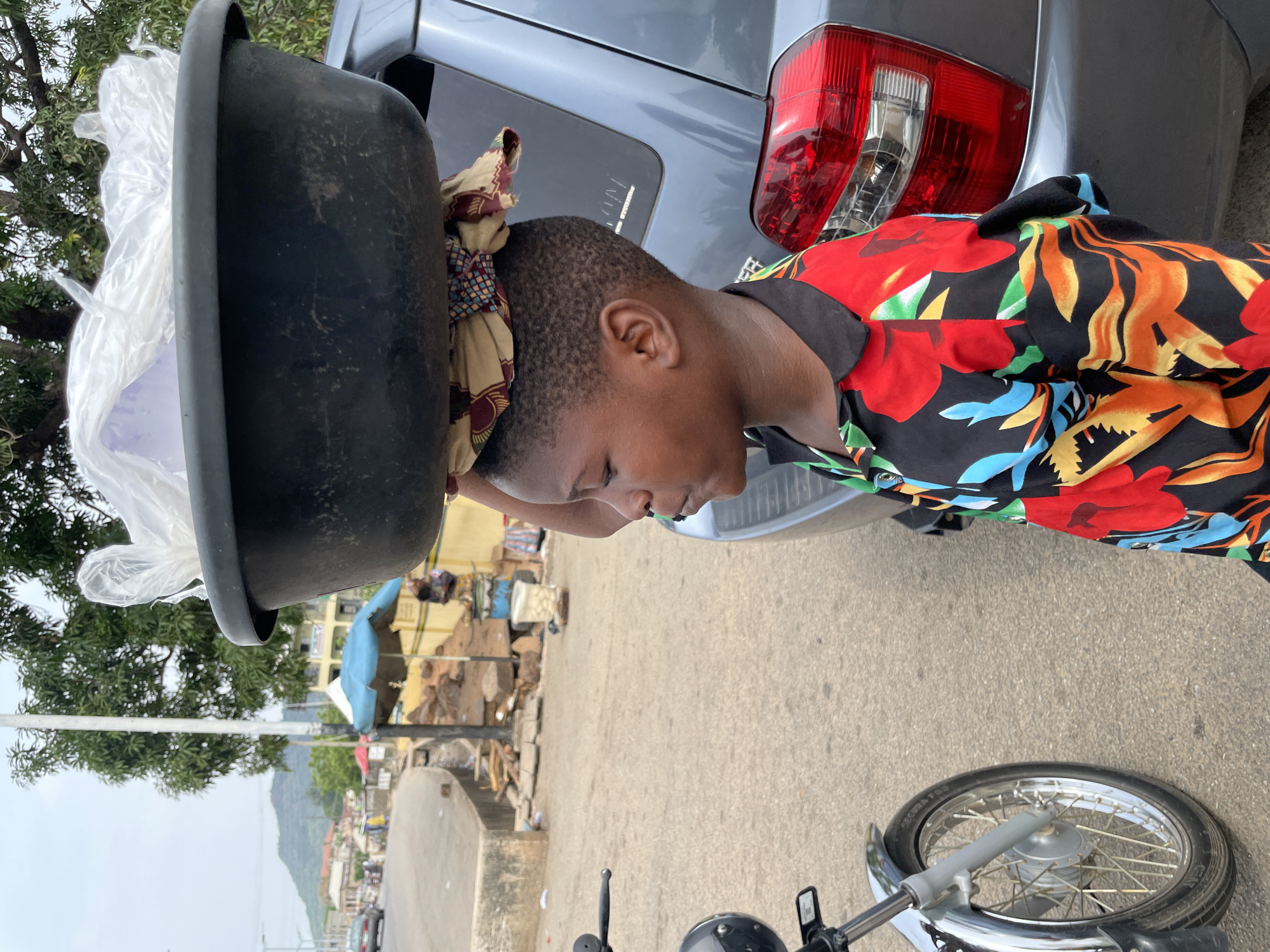 A young person, likely female, balances a large black basin on their head while standing next to a gray car and a motorbike. The basin is partially filled with what appears to be plastic bags. The individual is wearing a vibrant, floral-patterned shirt. The scene appears to be a street in Lokoja, Nigeria, characterized by a mix of low-rise buildings and local infrastructure. In the background, there's a street scene with various structures, including buildings with blue tarps. A tree is visible to the left, and in the distance, a mountain range can be observed. The motorbike's front wheel is visible in the foreground. The photograph is taken at a low angle, and the image is slightly rotated.