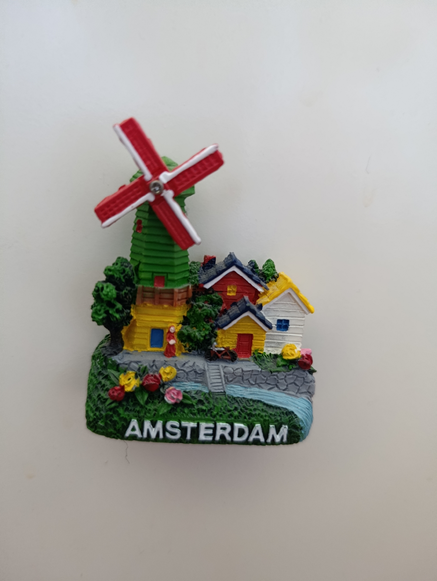 This is a close-up, eye-level, vertical, studio shot of a novelty souvenir, likely a refrigerator magnet, designed to represent a scene from Amsterdam. The souvenir features a miniature windmill with red and white blades set against a backdrop of small, colorful houses with red, yellow, and white roofs. The windmill is green and yellow. Lush green trees, red and yellow flowers, and a blue, water-like feature with a stone embankment are also depicted. The word "AMSTERDAM" is written in white, block letters across the front of the base. The background is a plain, light-colored, textured surface, possibly a wall. The lighting is even, with no harsh shadows. There are no people or animals visible in the scene. The souvenir is positioned in the center of the frame.