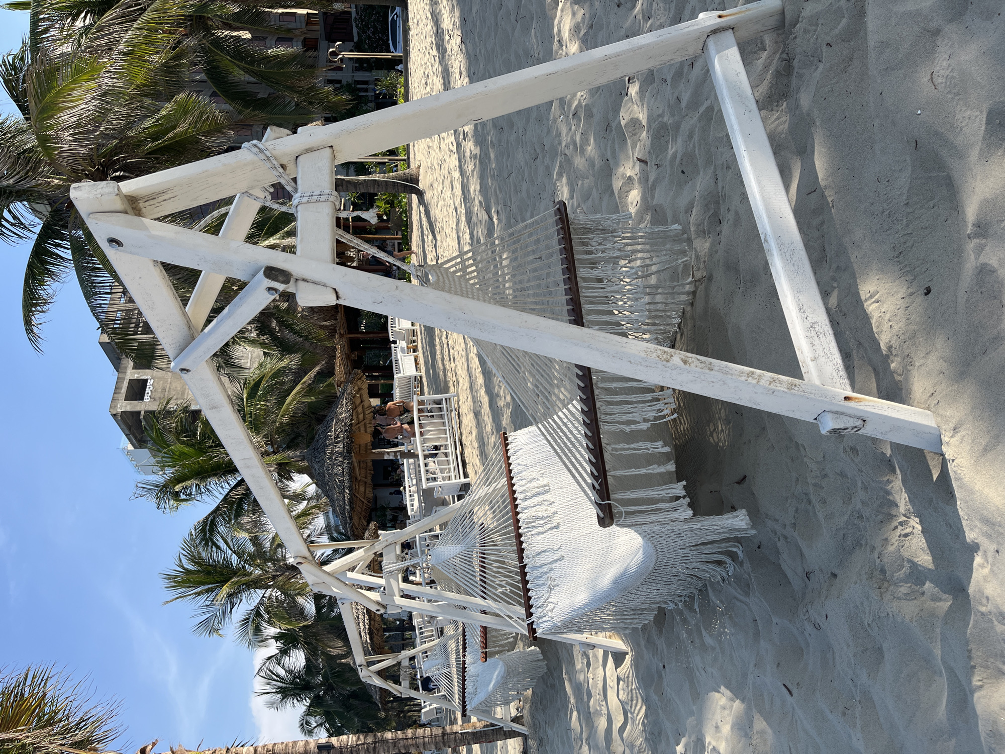 The image depicts a white hammock stand on a sandy beach. The hammock itself is also white with a fringed edge. Palm trees are visible in the background, along with a building under construction. There is also a thatched-roof structure with tables and chairs nearby where people are sitting. The sky is blue and clear. The image was most likely taken in the city of Đà Nẵng, Vietnam because that is the location specified in the request.