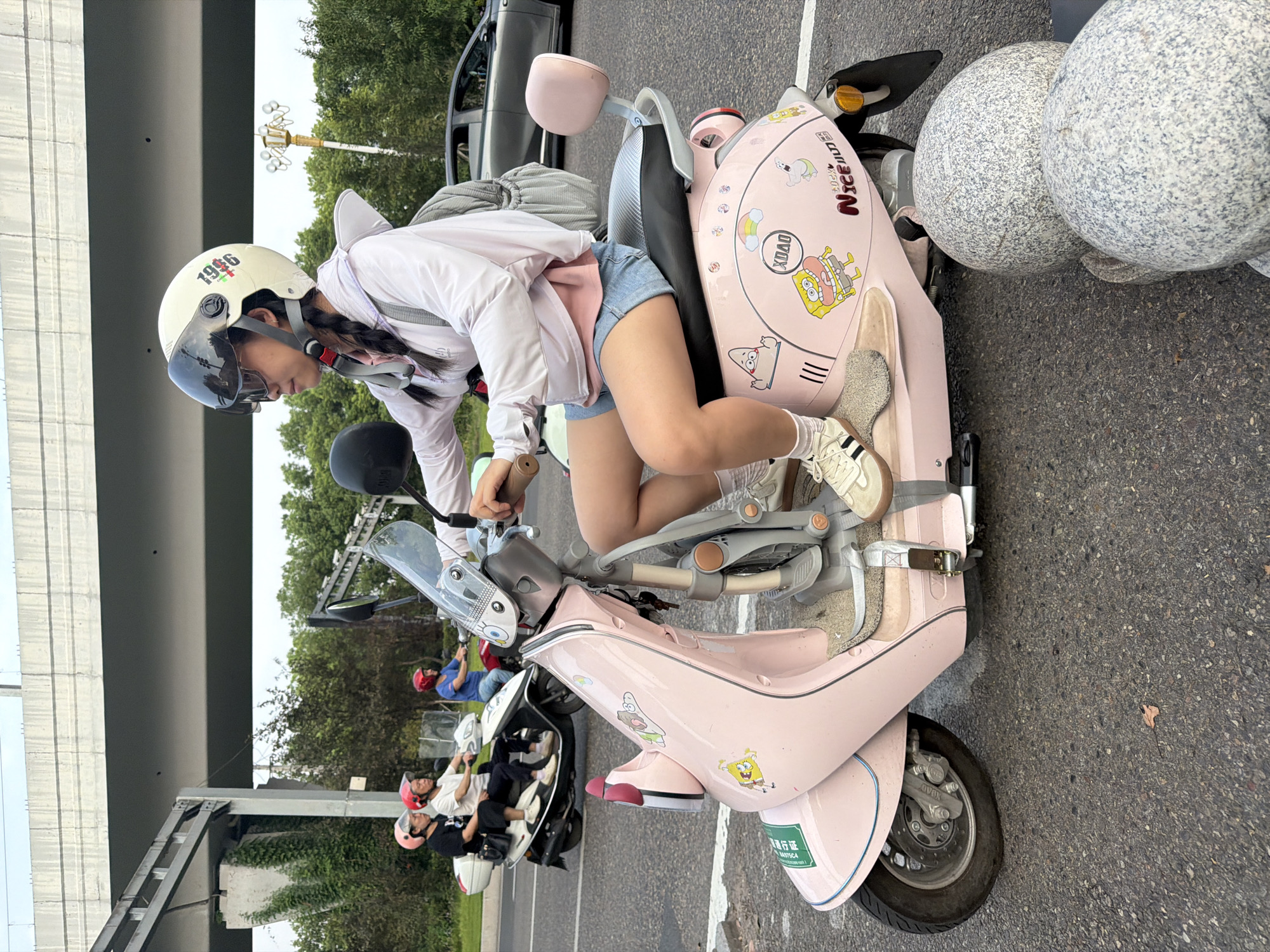 A young Asian woman with braided hair wearing a white helmet, sunglasses, a pink hoodie over a pink top, and denim shorts rides a pink scooter on a paved road in Jiangjin, China. She is wearing white sneakers with black stripes and white socks. The scooter is decorated with stickers of cartoon characters, including SpongeBob SquarePants and Patrick Star, and a rainbow. She is looking to her left. In the background, other people are riding scooters, and a gray car is visible. An elevated concrete structure and trees are also present in the scene. The road has white lines and is in good condition. The sky is overcast.