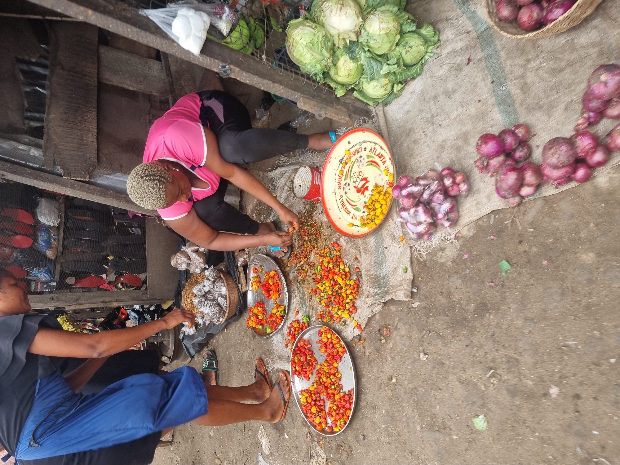 This image captures a vibrant and active outdoor market scene in Fegge, Nigeria, during the daytime, viewed from a high angle.
Two women are the primary human subjects, engaged in market activities. In the central foreground, a woman with short, light-colored hair, dressed in a pink top and dark leggings, is seated on the ground. She is meticulously processing a large quantity of colorful peppers, appearing to be stemming or de-seeding them. These peppers, a mix of red, orange, and yellow hues, are spread across a cloth on the ground and gathered into two oval metal trays and one large, round decorative tray.
To her left, another woman, wearing a black top and blue shorts, is also seated on the ground. Her hands are near a woven basket filled with individually wrapped items in clear plastic, which could be dried goods or local produce like mushrooms. Both women are wearing flip-flops or sandals.
The market stall surrounding them displays a variety of fresh produce. To the upper right, several heads of green cabbages are stacked on a wooden surface. Further to the right, numerous red onions are arranged in a woven basket and scattered on the ground. The background reveals rustic wooden structures and shelving containing other merchandise, possibly footwear or various packaged goods, indicating a diverse marketplace.
The ground is a rough, uneven surface, consistent with an outdoor market. Visible text on the large round tray includes "ATLANTA CONGRES" and "GOODS PRODUCT OF NIGERIA," offering a clear location cue. The overall atmosphere suggests industrious daily commerce in a local Nigerian market.
