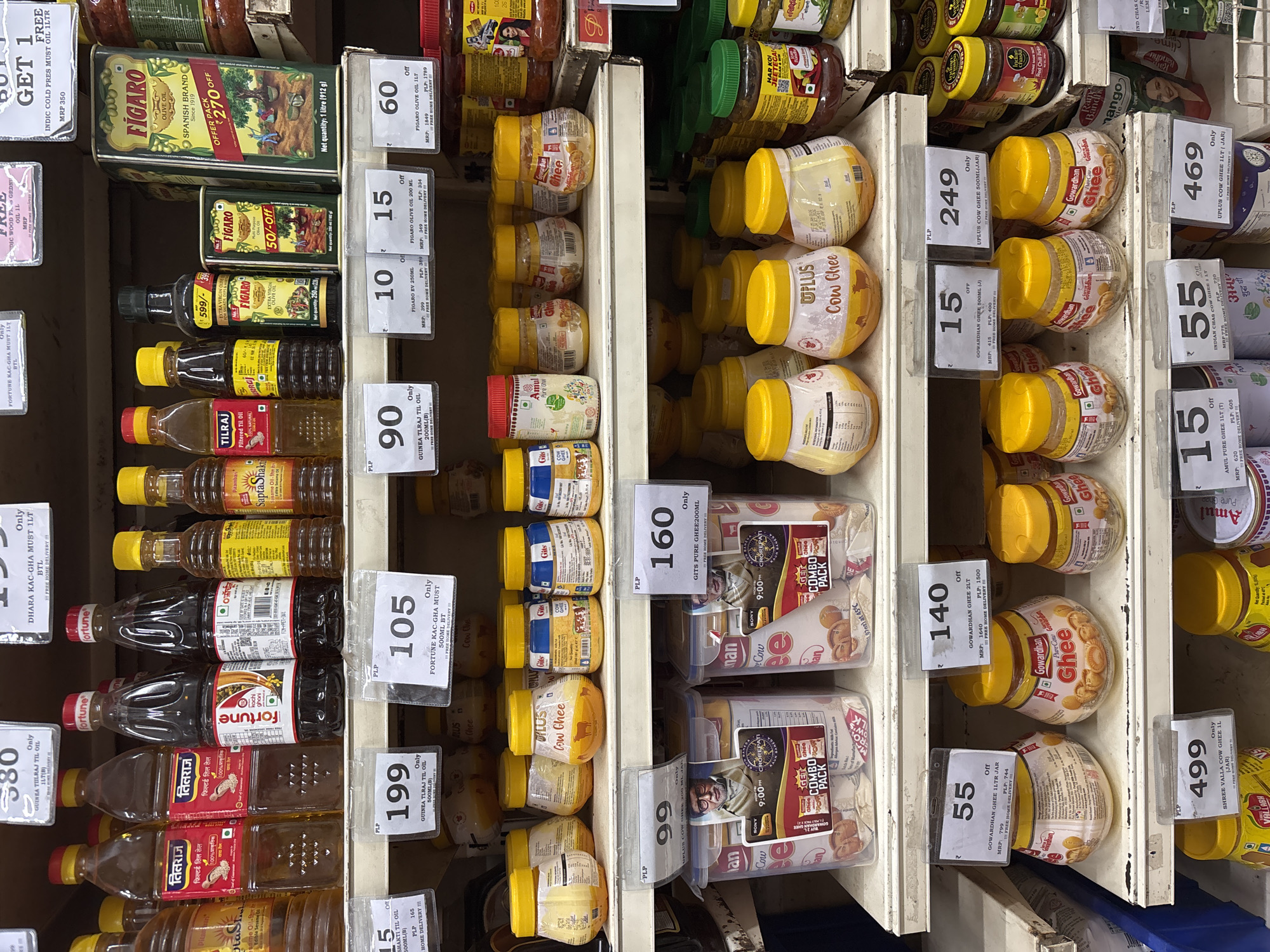 The image shows a shelf display in a store, presumably in Ambernath, India, filled with various brands of cooking oils and ghee.
On the left, there are multiple rows of cooking oils in plastic bottles. Brands such as "Tilraj," "Fortune," and "Saptashakti" are visible, with price tags ranging from ₹15 to ₹199. Some oils advertise being "filtered" or "100% natural." There are also promotions like "Buy 1 Get 1 Free."
The center and right of the image feature different ghee brands in jars, like "Uplus Cow Ghee," "Gowardhan Ghee," "Gits Pure Ghee," and "Amul Pure Ghee." Price tags range from ₹15 to ₹499. Some ghee products are advertised as "pure cow ghee." There are combo packs of "Gits" ghee and promotions on free home delivery.
The shelf itself appears to be a standard metal shelving unit, common in retail environments. The store is selling a variety of cooking products.