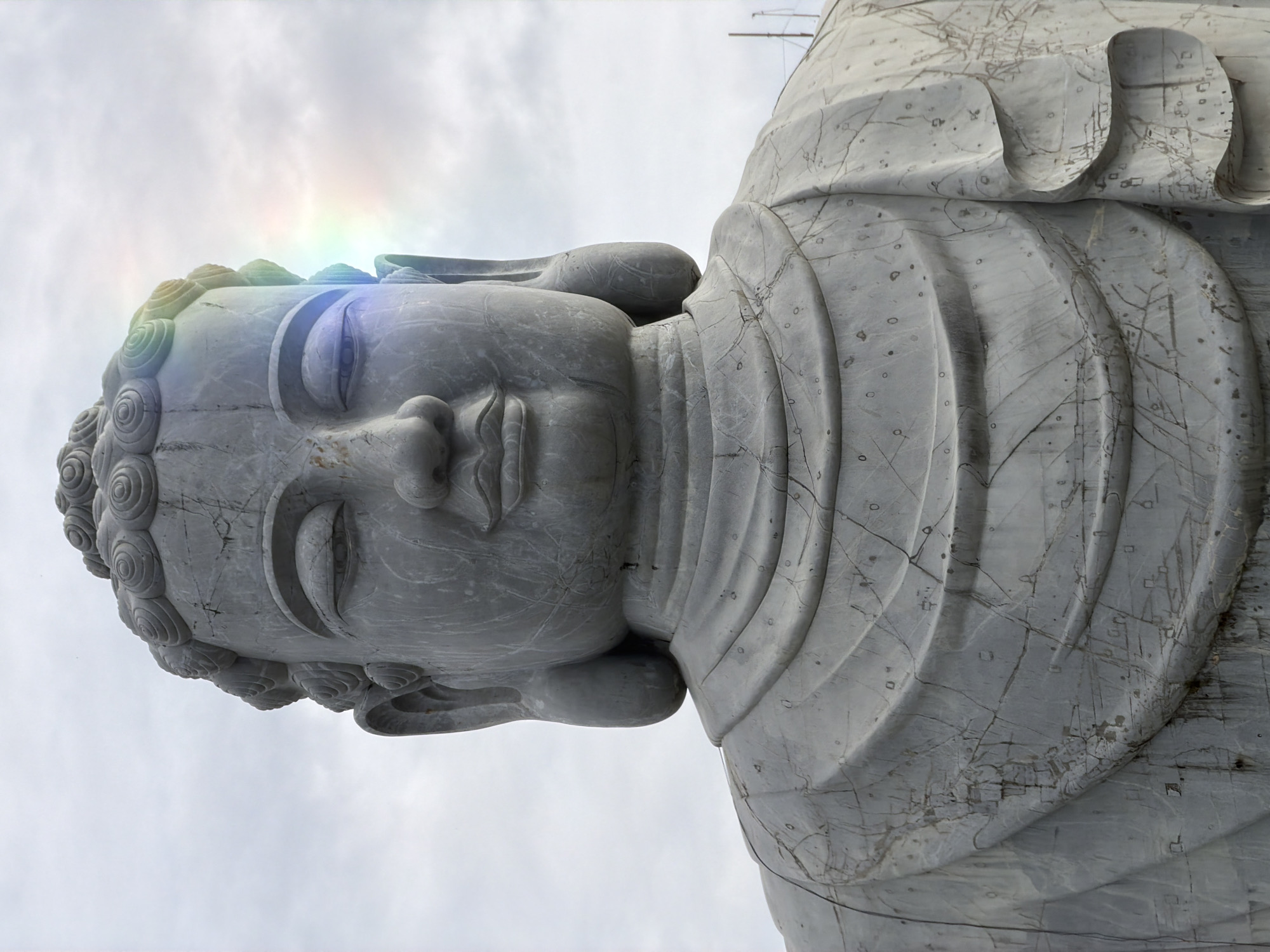 The image shows a close-up, upward-facing view of a gray stone Buddha statue against a cloudy sky. A faint rainbow-like effect appears on the statue's head. The statue exhibits carved details including the hair and draped clothing. The stone surface shows signs of weathering and wear. This statue is located in the city of Đà Nẵng, Vietnam.