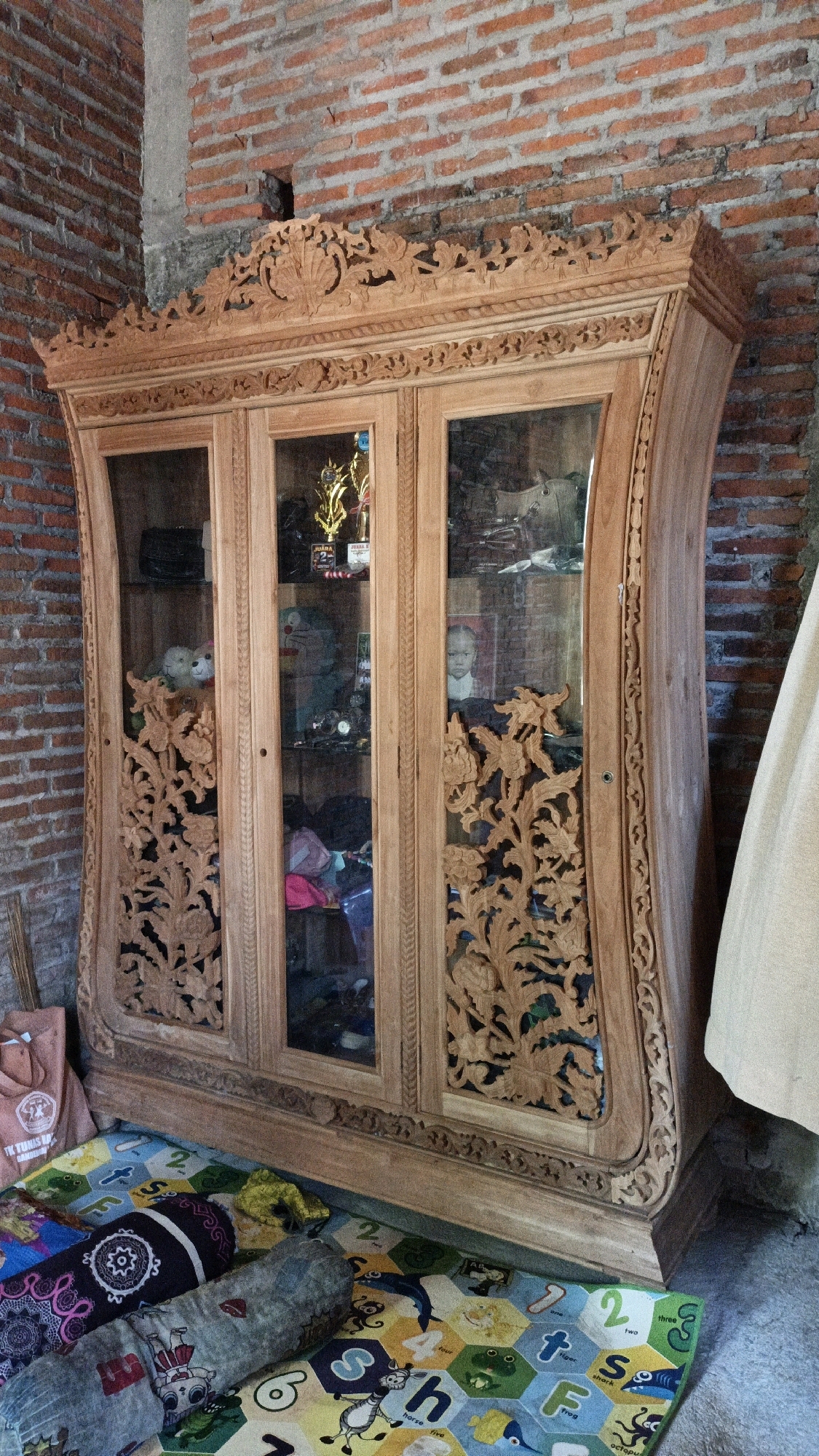 The image shows a wooden display cabinet in what appears to be a home or storage space. The cabinet is intricately carved with floral motifs and has glass doors, revealing various items inside, including trophies, stuffed animals, and other objects. The cabinet is positioned against a brick wall and stands on a colorful mat with alphabet and animal designs. In the foreground, there are also some rolled-up items and a pink t-shirt. The environment seems to be in Cikal, Indonesia, based on the description.