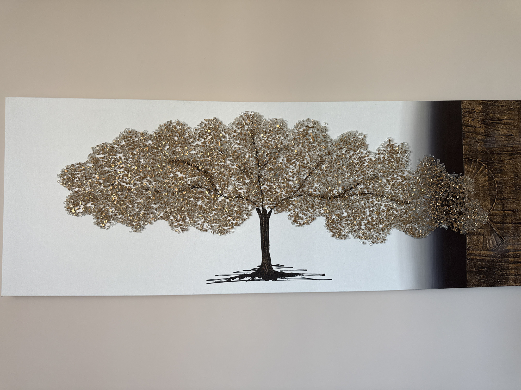 The image shows a horizontal art piece depicting a tree with a full, rounded canopy made of small, shimmering golden-brown elements, possibly crystals or beads, against a white background. The tree trunk and roots are illustrated in black ink. The right edge of the artwork transitions into a vertical band of gradated black and a smaller panel with a textured, golden-brown design featuring radiating lines, adding depth to the composition. The artwork is mounted on a neutral-colored wall. The location is Unknown, Unknown.