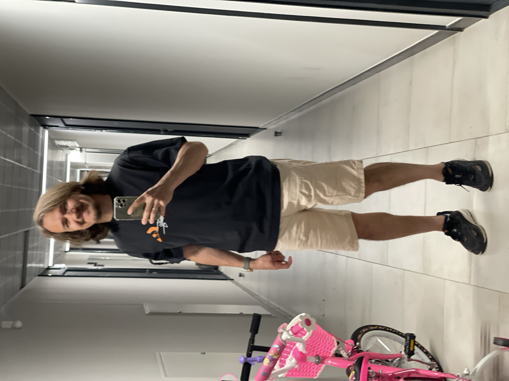 A man with shoulder-length brown hair and a beard is depicted taking a mirror selfie in an indoor hallway, rotated 90 degrees counter-clockwise. He is smiling and looking towards his phone, which he holds up to capture his reflection. He is wearing a black t-shirt with a partially visible graphic design, possibly displaying "Stussy" and an orange motif. He also wears light beige shorts and dark athletic shoes with white soles and white socks. A silver-colored watch is on his left wrist, and a ring is on his right ring finger.
The setting appears to be a modern hallway with light-colored walls and floor, possibly tiled. The ceiling is white with dark trim and integrated lighting. Dark doors are visible in the background along the hallway. In the foreground, lying on its side, is a bright pink child's bicycle with a white basket, white tires, and colorful decorations, including purple elements on the handlebars. The overall lighting suggests an indoor, artificial light source. The man's expression is pleasant and relaxed.