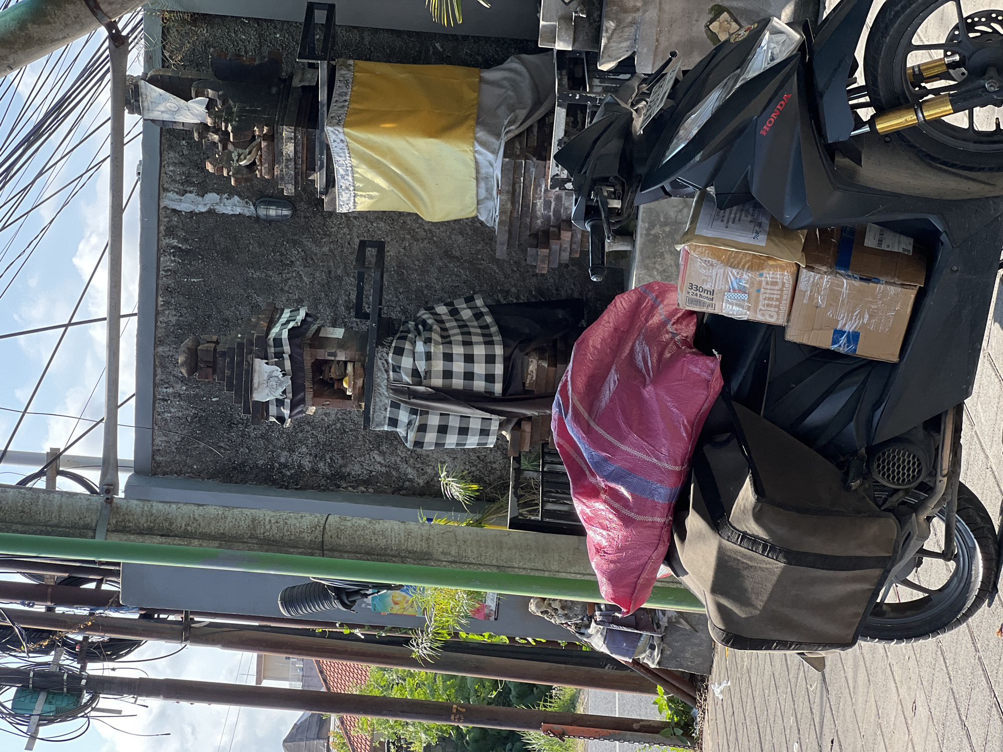 A black Honda scooter is parked on a tiled sidewalk in Denpasar, Indonesia, loaded with packages and a large pink plastic-wrapped bundle. The scooter has brown saddlebags mounted on the rear. To the right of the scooter, there is a stone shrine adorned with yellow and white fabric. Behind the scooter and shrine is a grey textured wall. In the background, to the left, a building with a red tile roof is visible amidst lush green foliage. Overhead, a dense network of electrical wires crisscrosses the sky, strung between utility poles. The scene is illuminated by bright daylight.